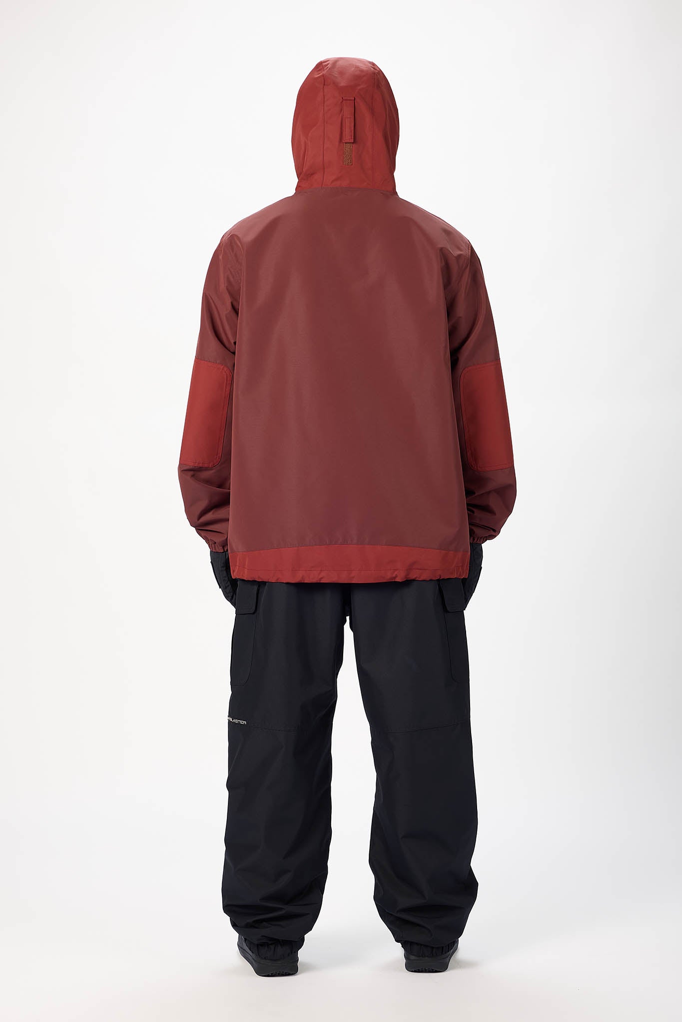A person shown from the back wearing a hooded jacket with a dark reddish-brown main body and darker red sleeves, paired with black pants, against a plain light gray studio background.