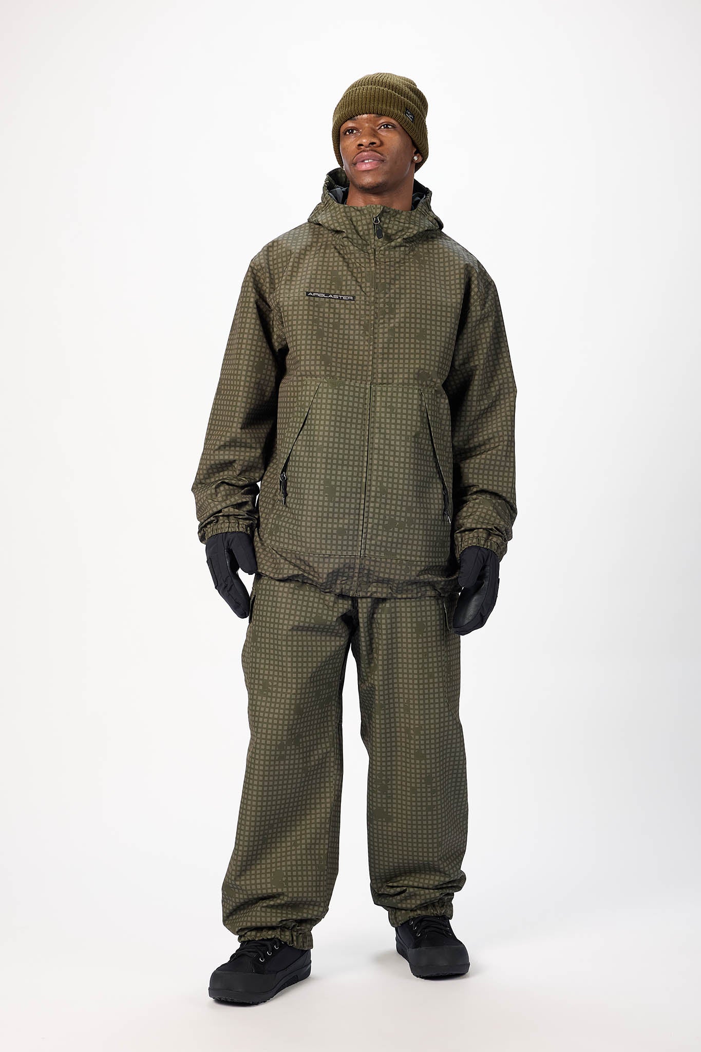A man modeling an olive green and black checkered hooded jacket and matching pants, paired with black gloves, a black beanie, and black snow shoes, against a plain white studio background.