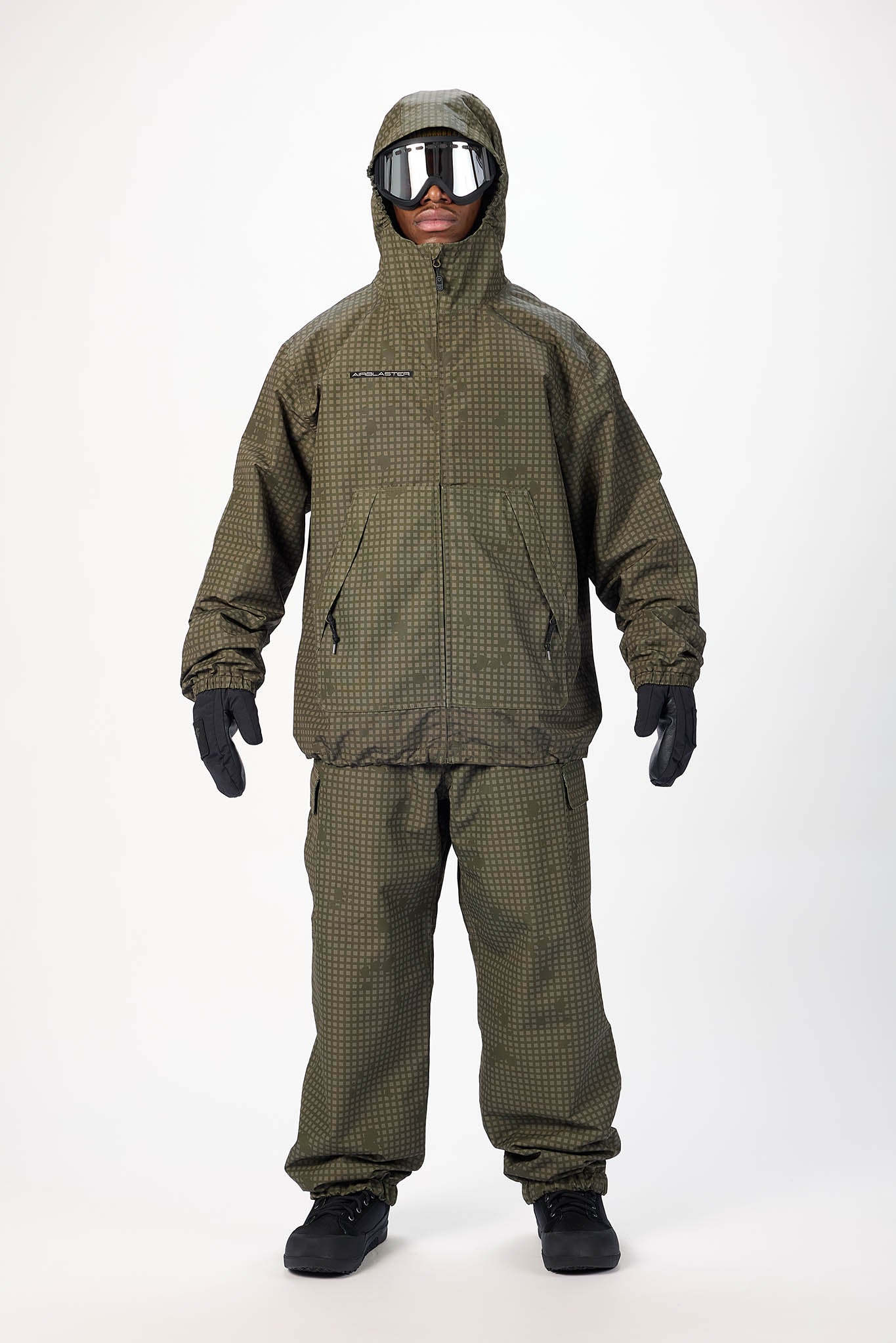 A person modeling a hooded snowsuit in an olive green and black micro-check pattern, paired with black gloves, goggles, and black shoes, against a plain white studio background.