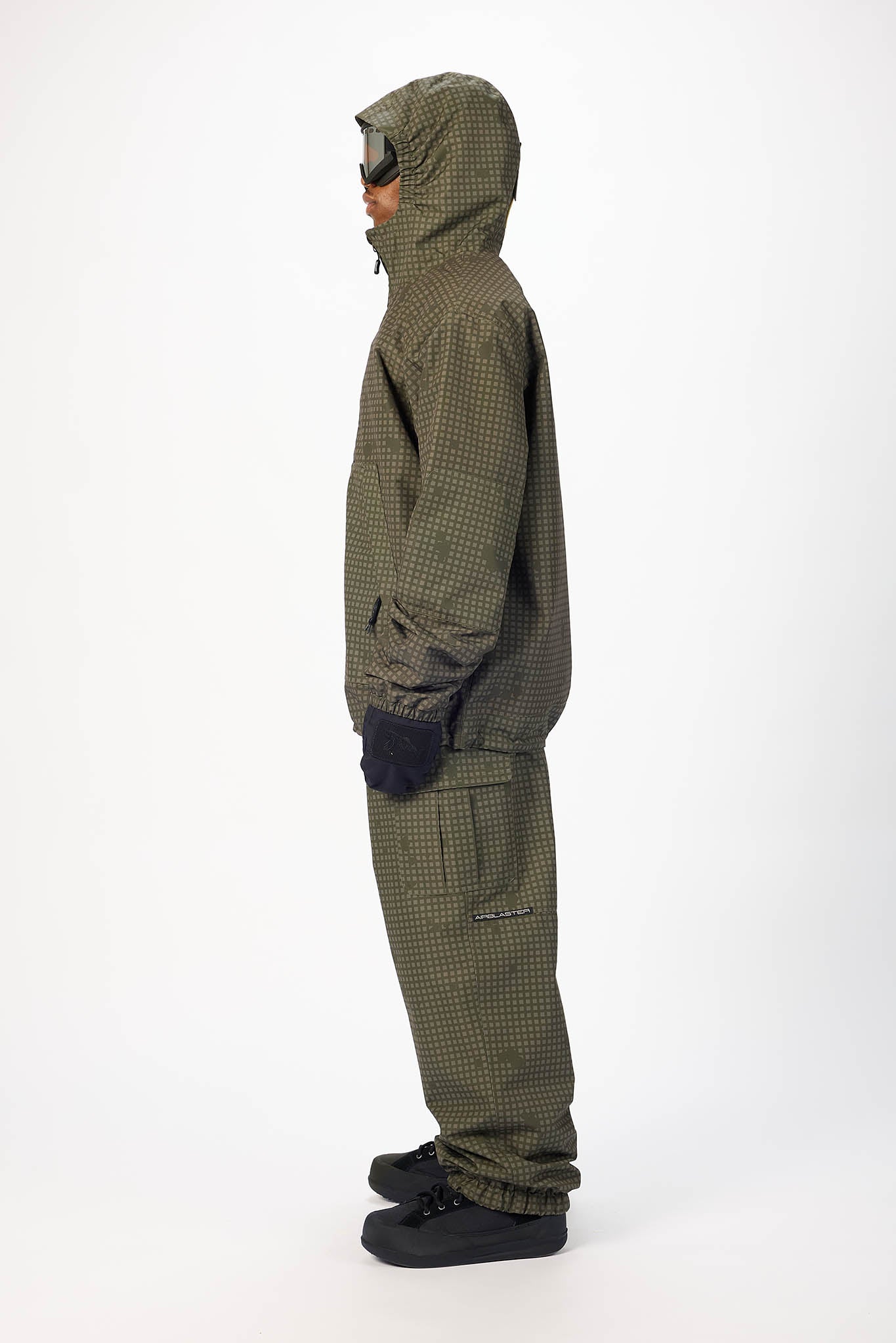 A person shown in profile wearing an olive green hooded jacket and matching pants with a small check pattern, black gloves, and black shoes against a plain white studio background.