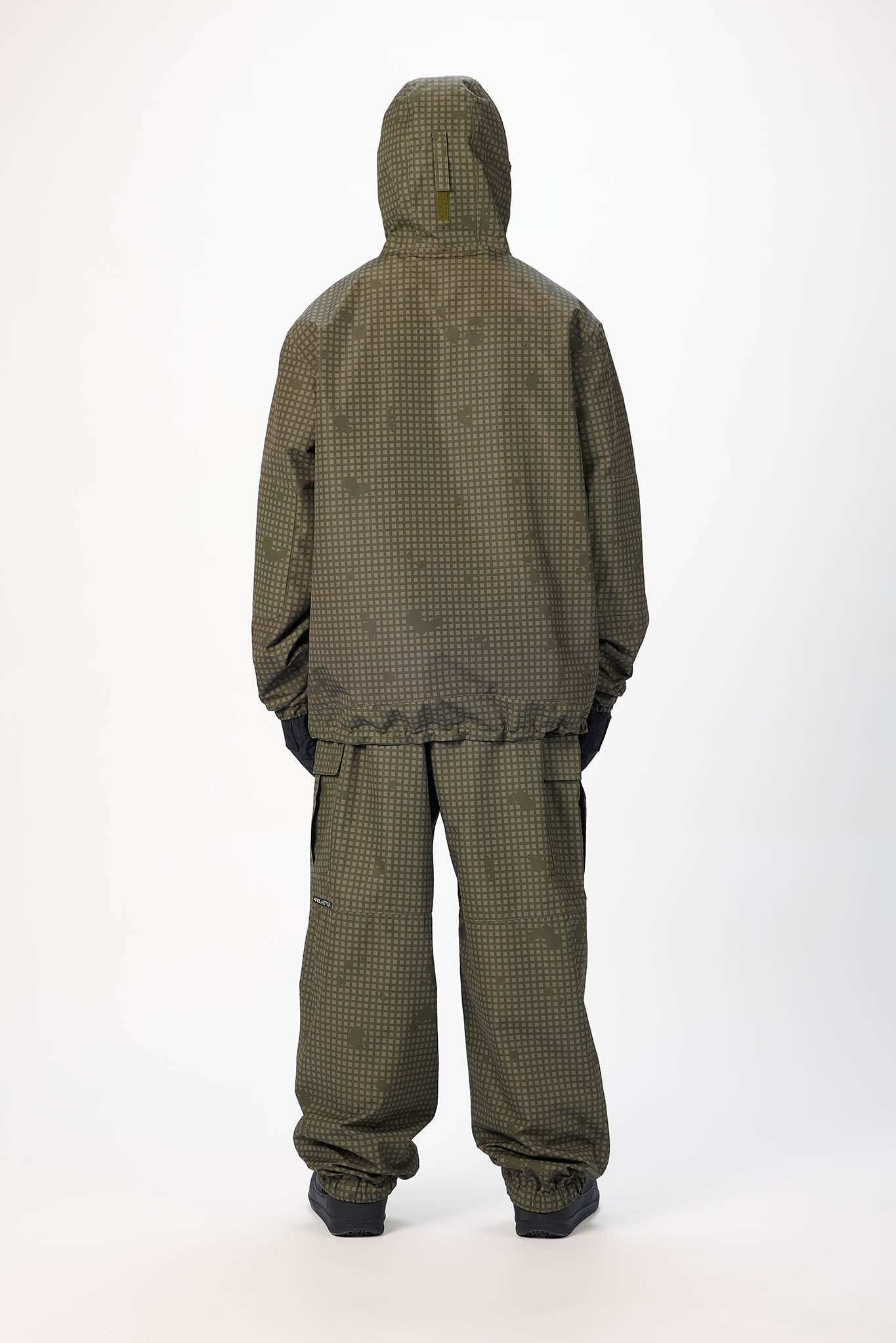 A person viewed from the back wearing an olive green hooded jacket and matching loose pants with a small check pattern, standing against a plain light gray studio background.