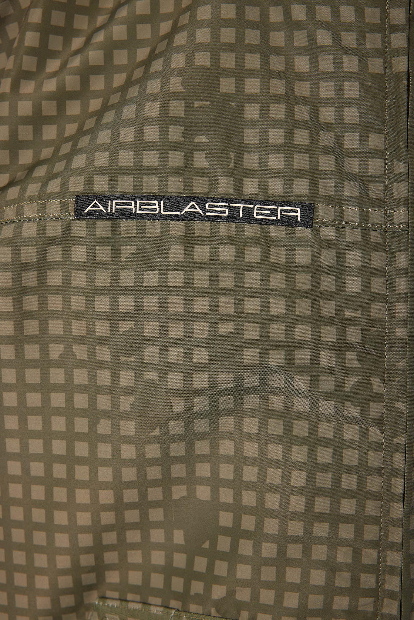 Close-up of a textured fabric with a beige and olive green grid pattern and a black label reading 'AIRBLASTER'.