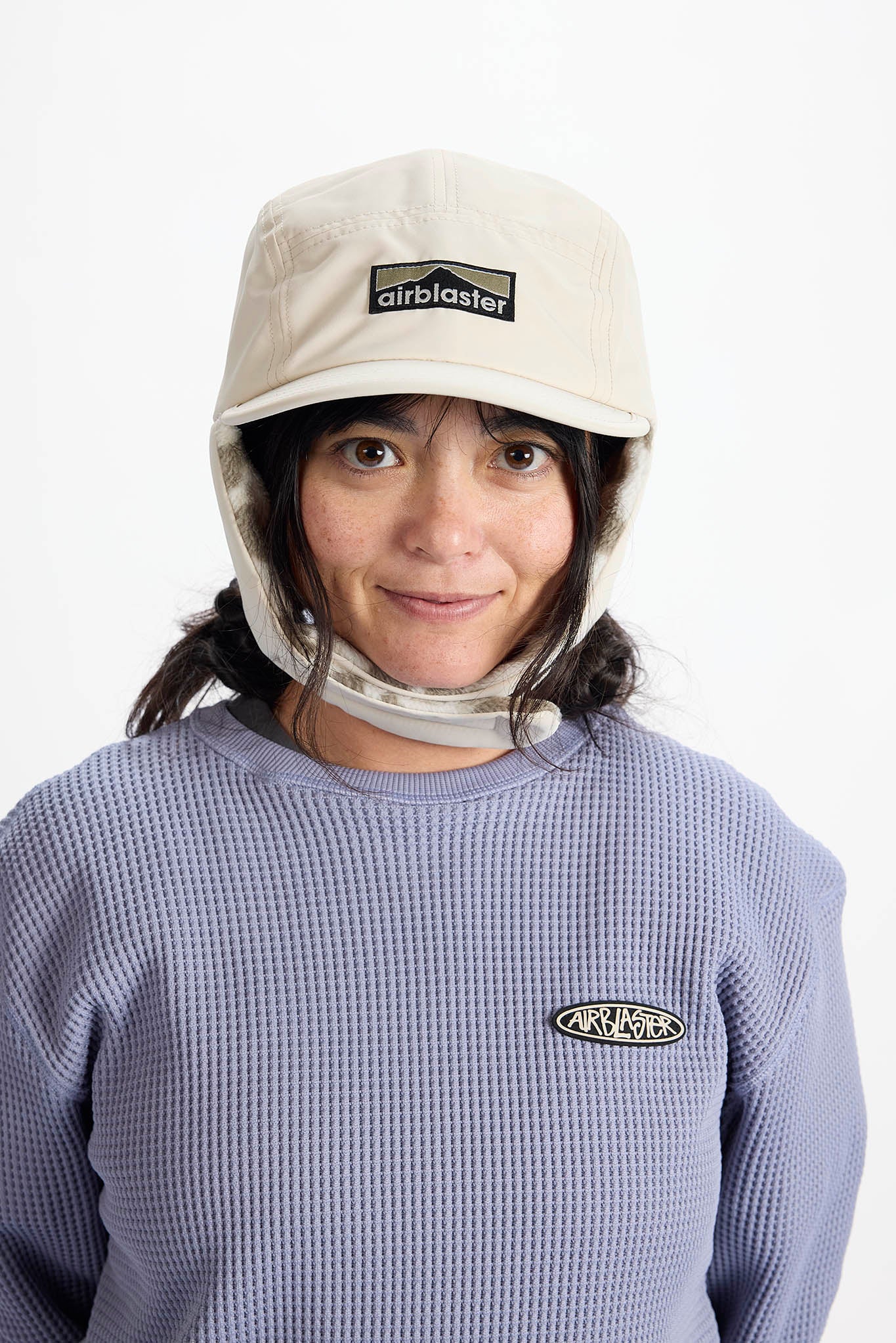 A woman wearing a light beige Airblaster cap with ear flaps and a clip on the bottom