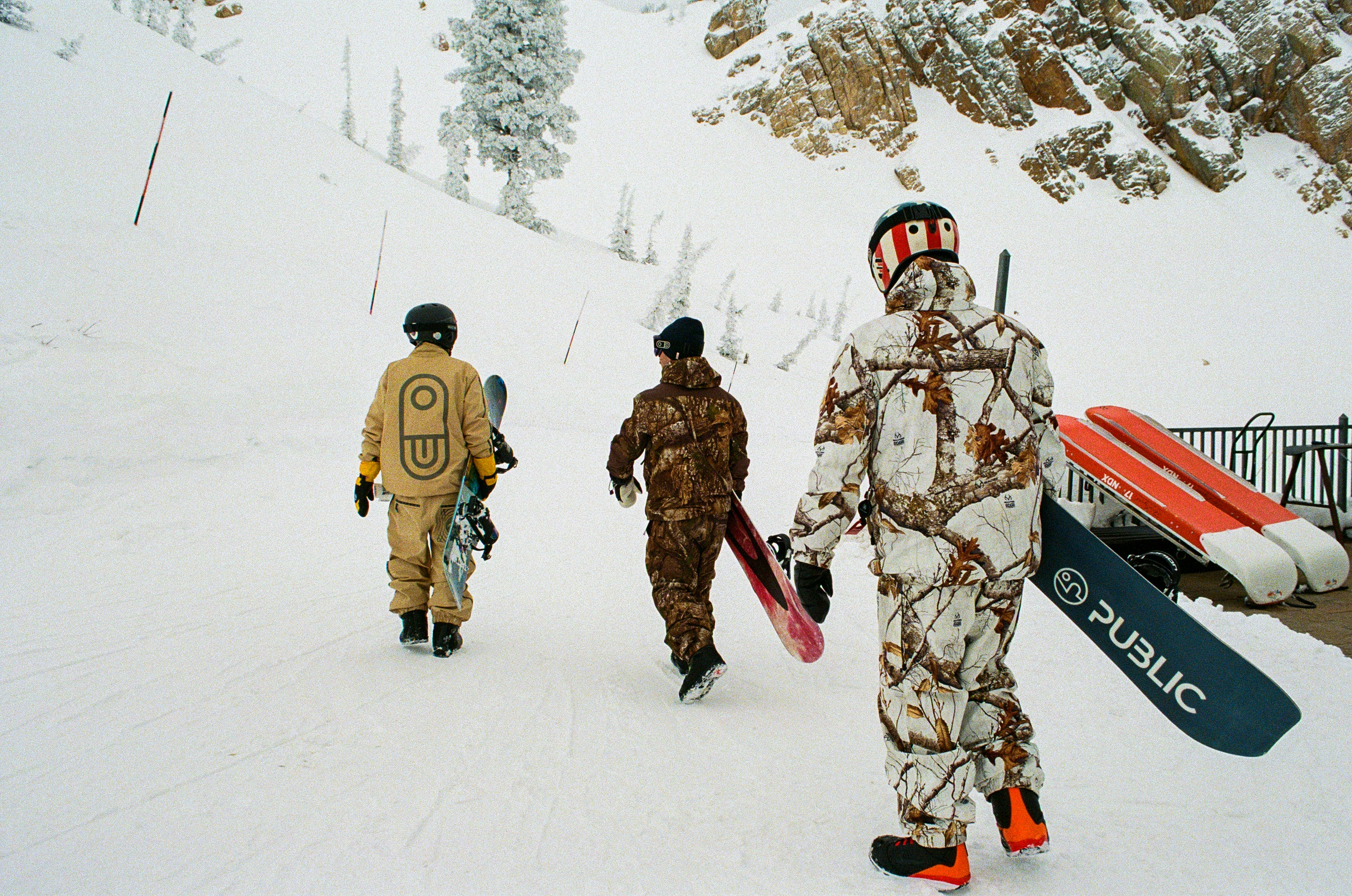 Three snowboarders walking down a snowy mountain slope, each carrying a snowboard. The person on the right wears a white camouflage Public snowsuit and carries a black Public snowboard.