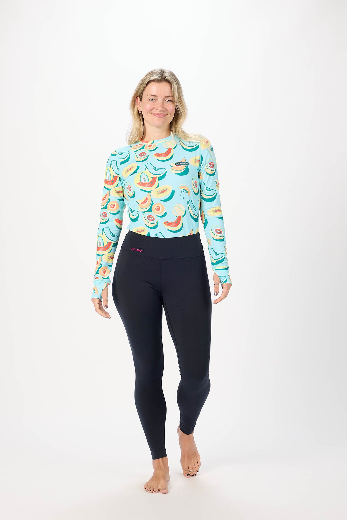 A woman modeling a long-sleeve athletic top with a colorful watermelon pattern on a light blue background, paired with black leggings, against a plain white studio background.