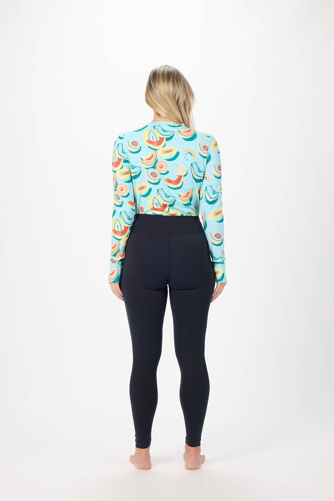 A woman viewed from behind wearing a light blue long-sleeve crop top with a colorful watermelon print and black leggings, standing against a plain white studio background.