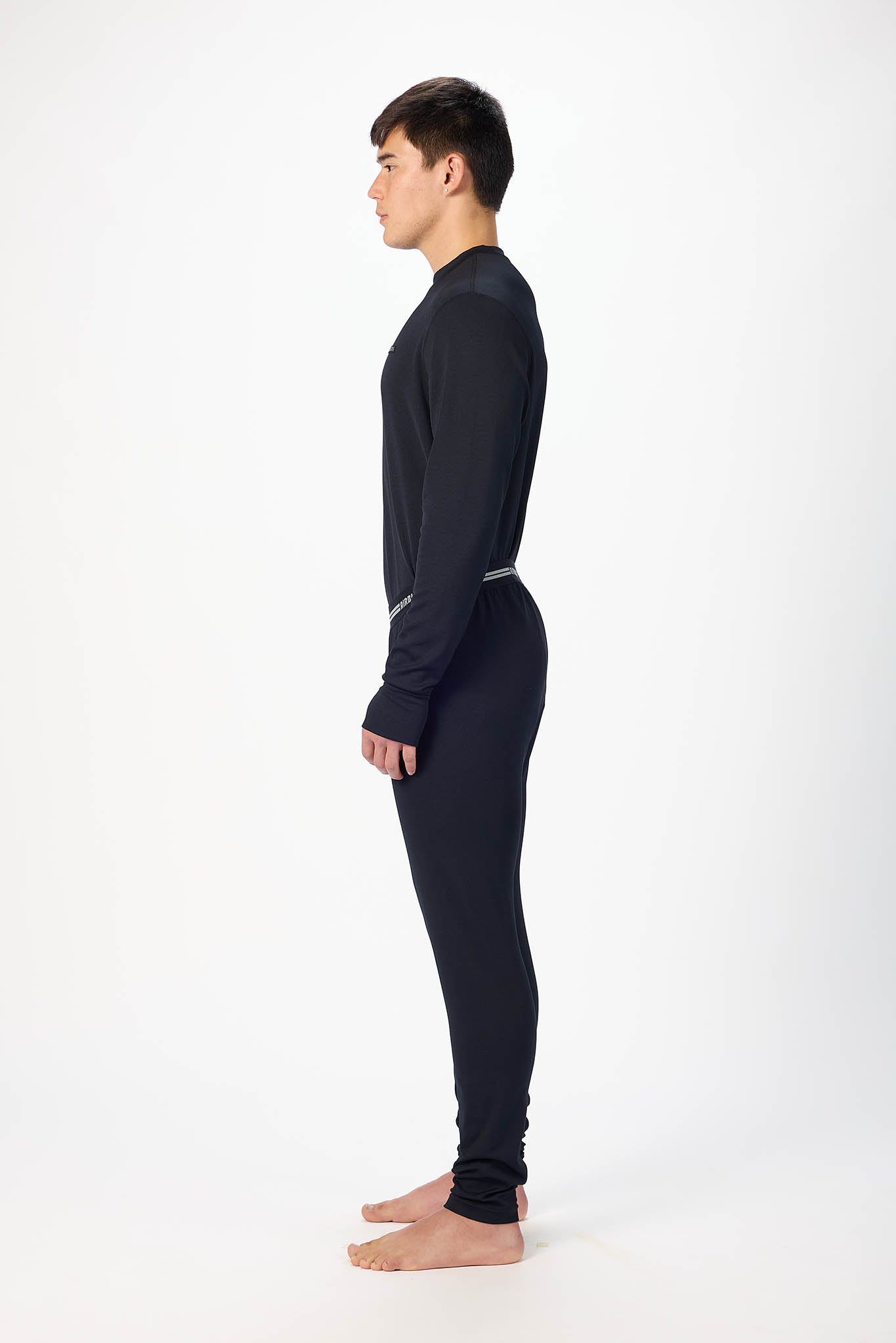 in side profile A man modeling a black long-sleeve crewneck base layer and matching black pants with a thin silver waistband, standing against a plain white studio background.