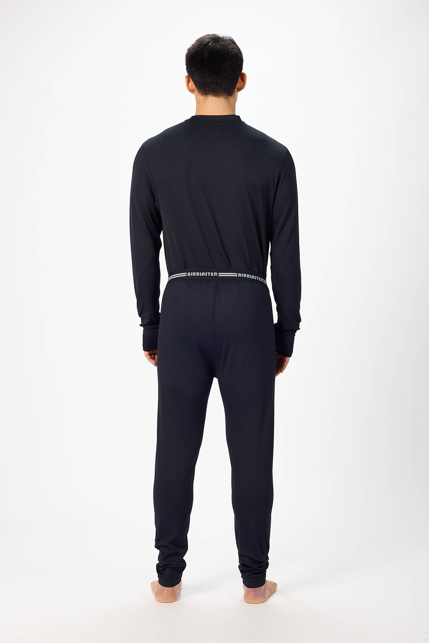 A man viewed from the back wearing a black long-sleeve top and matching black pants with a thin silver stripe detail at the waist, standing against a plain white studio background.