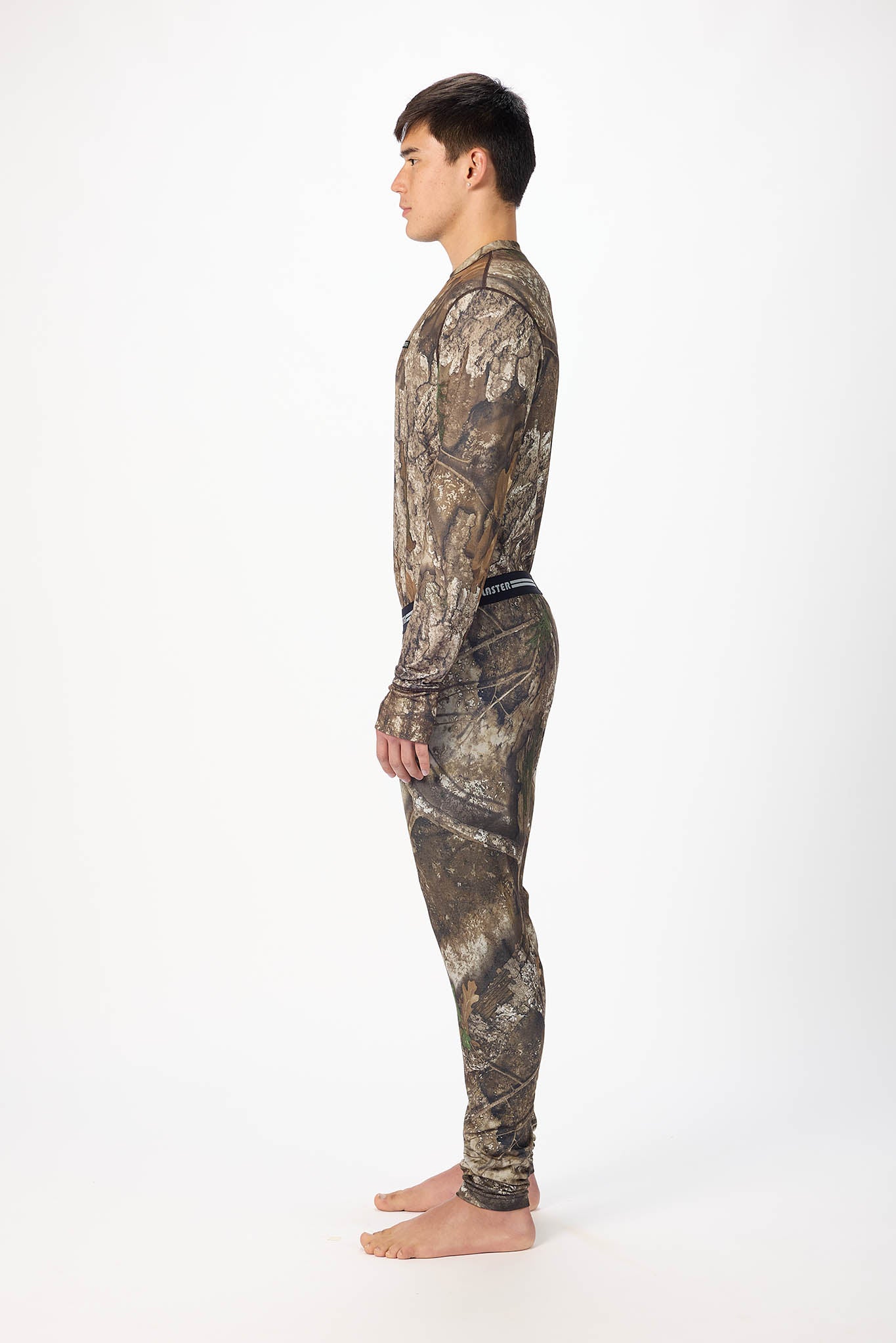 A man in profile wearing a long-sleeve camouflage shirt and matching camouflage pants, standing barefoot against a plain white studio background.