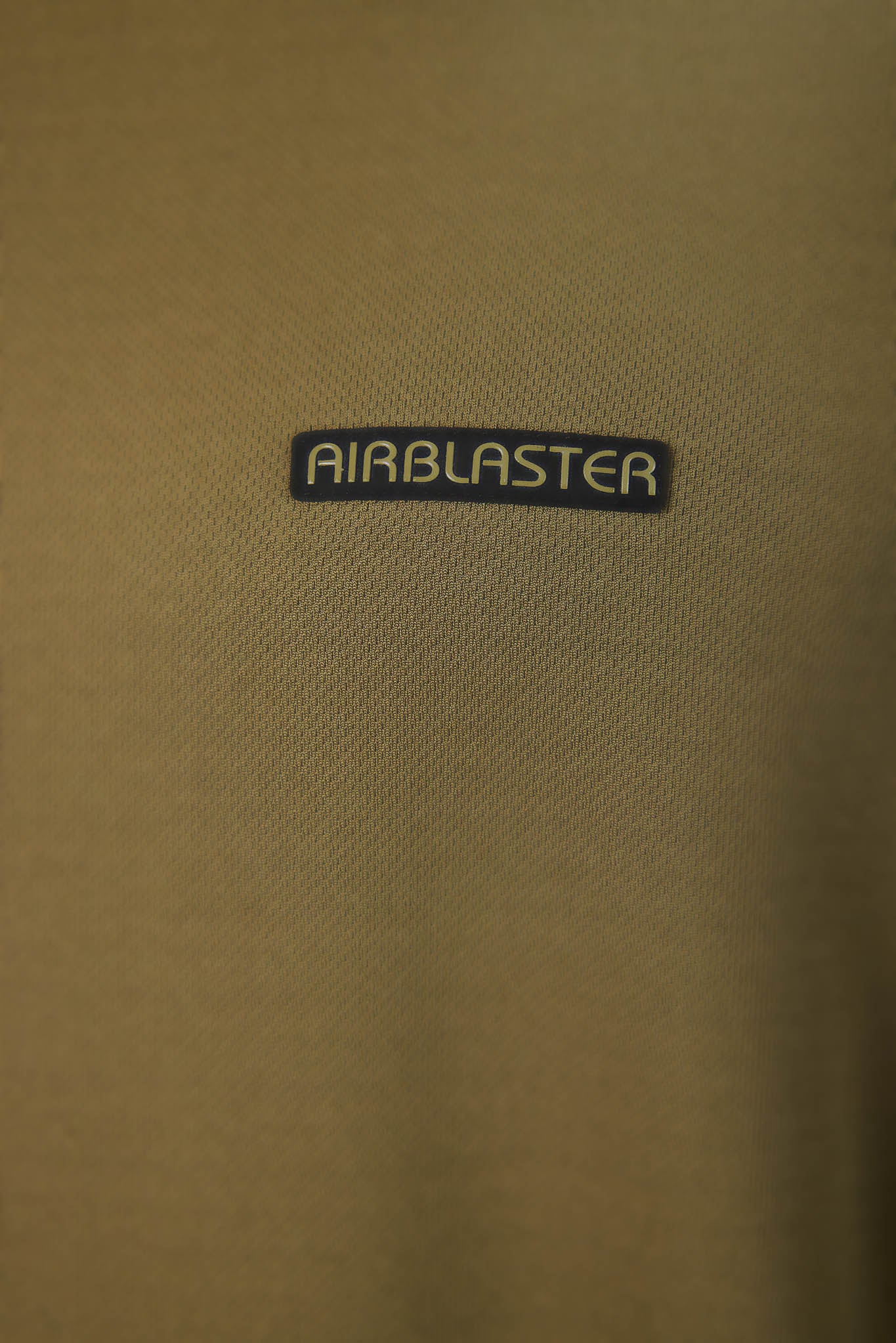 Close-up of a tan textured fabric with a small black rectangular patch reading AIRBLASTER in white letters.