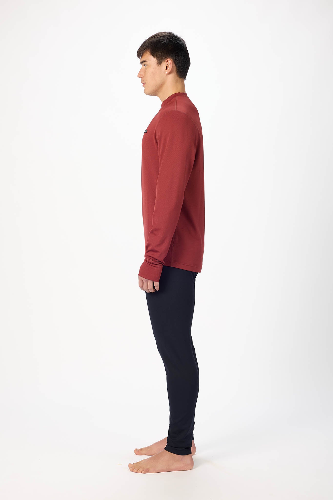 A man shown in profile standing barefoot against a plain white studio background, wearing a long-sleeve crew-neck top in muted brick red and dark navy pants.