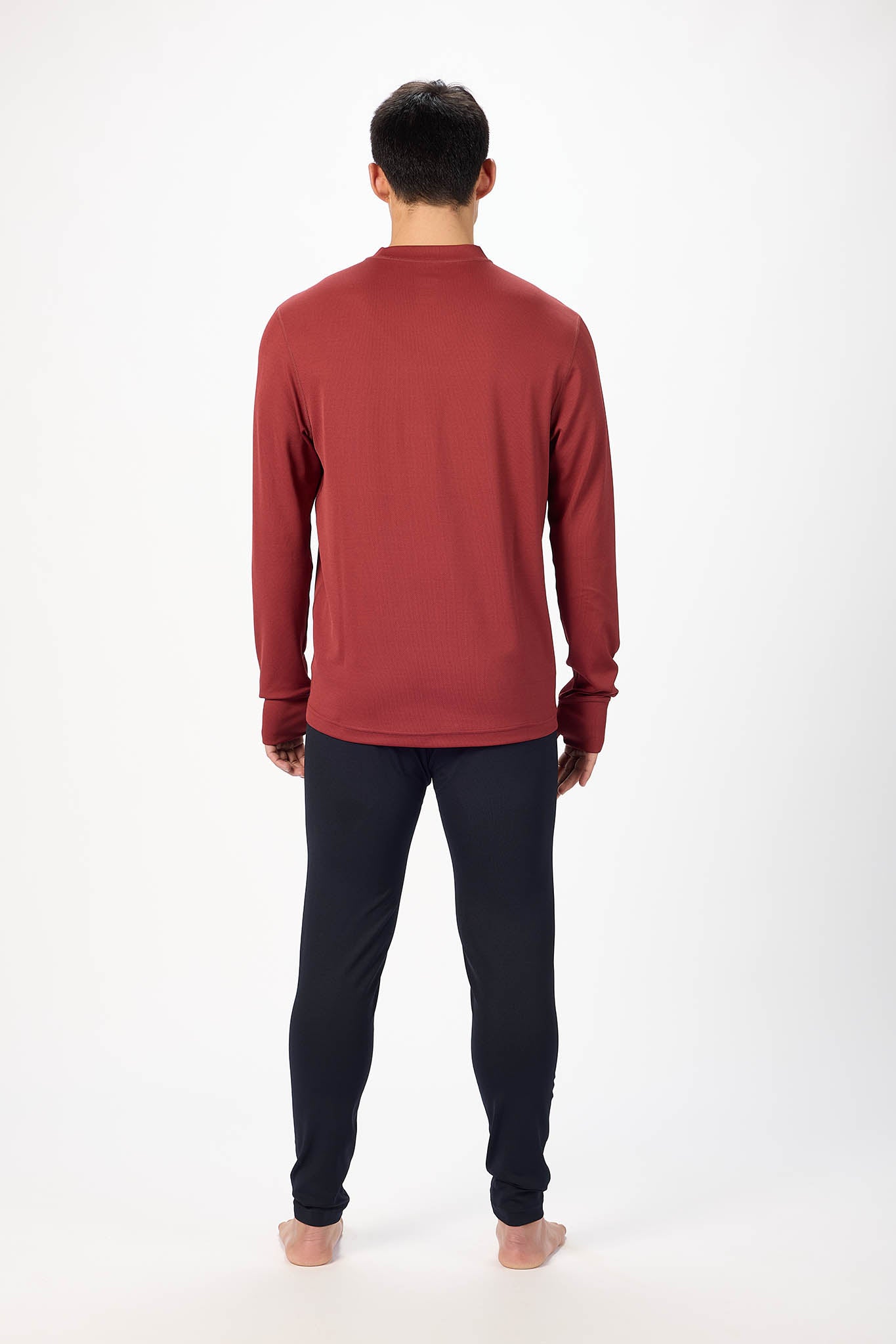 A man viewed from the back wearing a long-sleeve crewneck shirt in a muted brick red color and dark navy pants, standing against a plain white studio background.