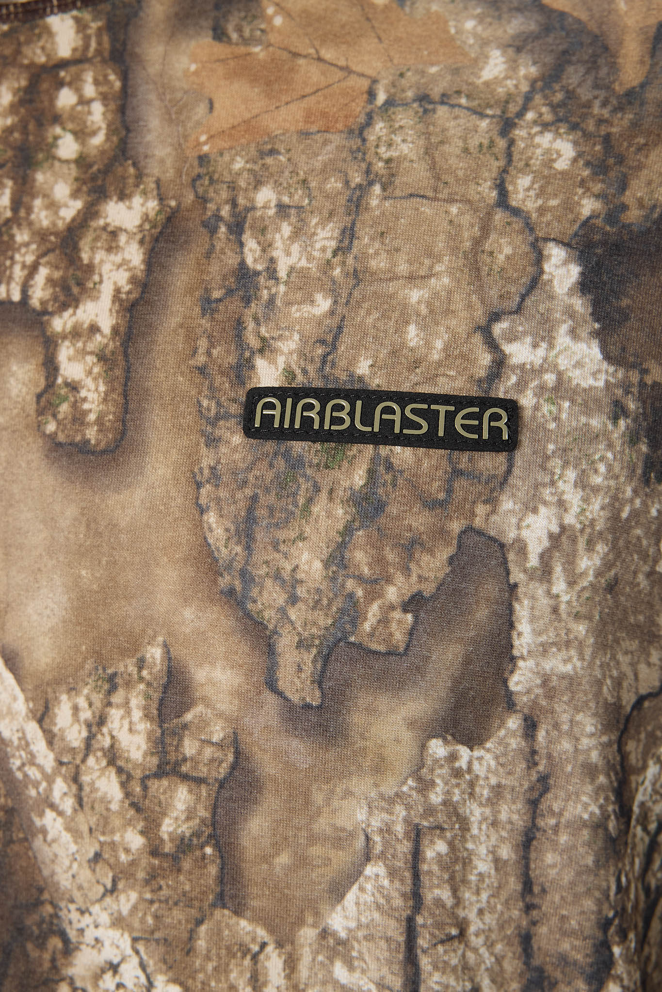 A close-up of a textured camouflage pattern in earthy brown, beige, and gray tones, featuring a black rectangular patch with the word 'AIRBLASTER' in light lettering.