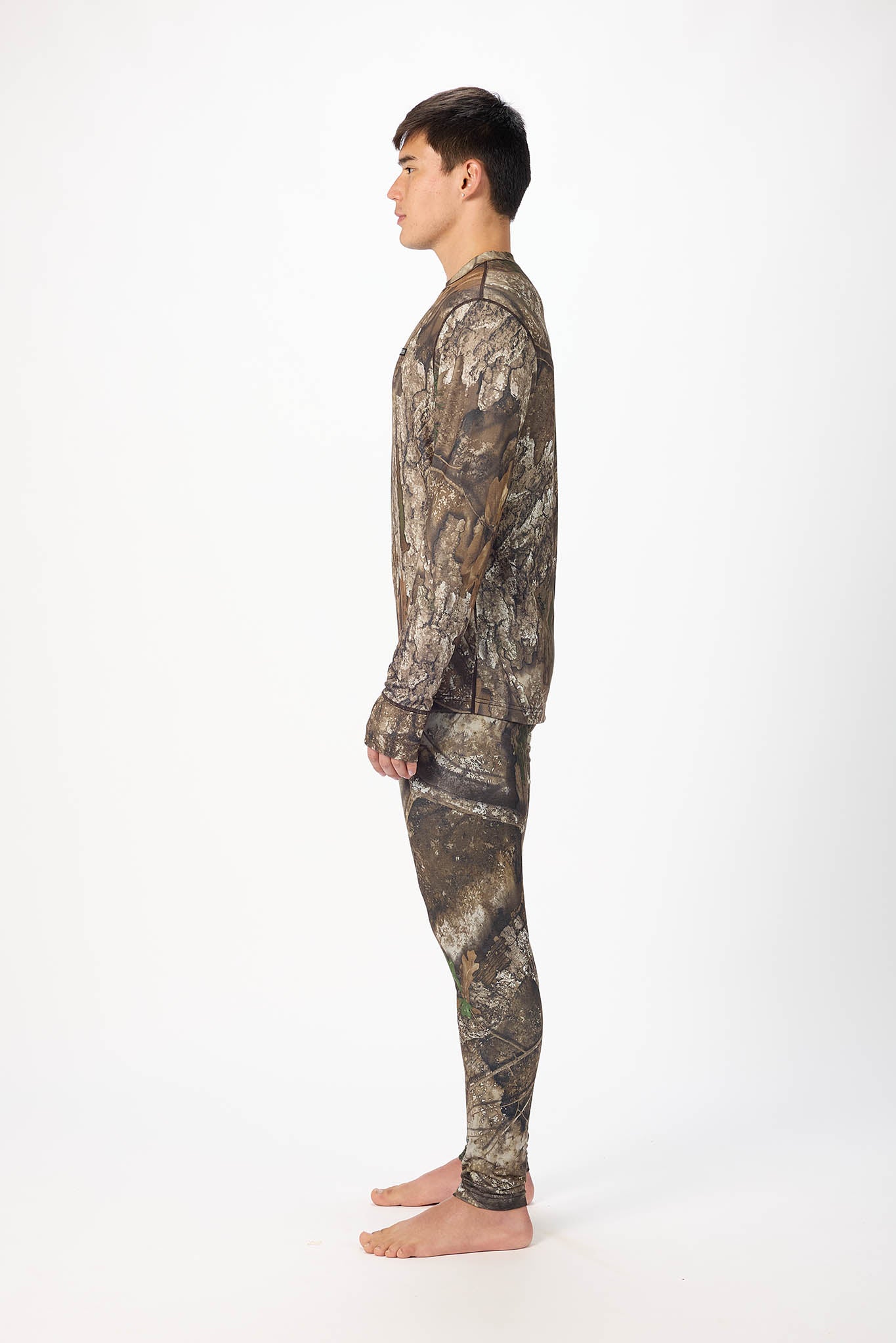 A man shown in profile wearing a long-sleeve camouflage shirt and matching camouflage pants, standing barefoot against a plain white studio background.