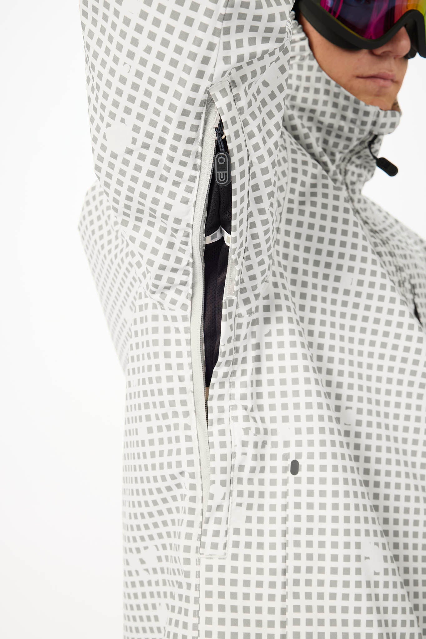 Close-up of a person wearing a white ski jacket with a gray square grid pattern, showing the hood, zipper, and part of the face with ski goggles.