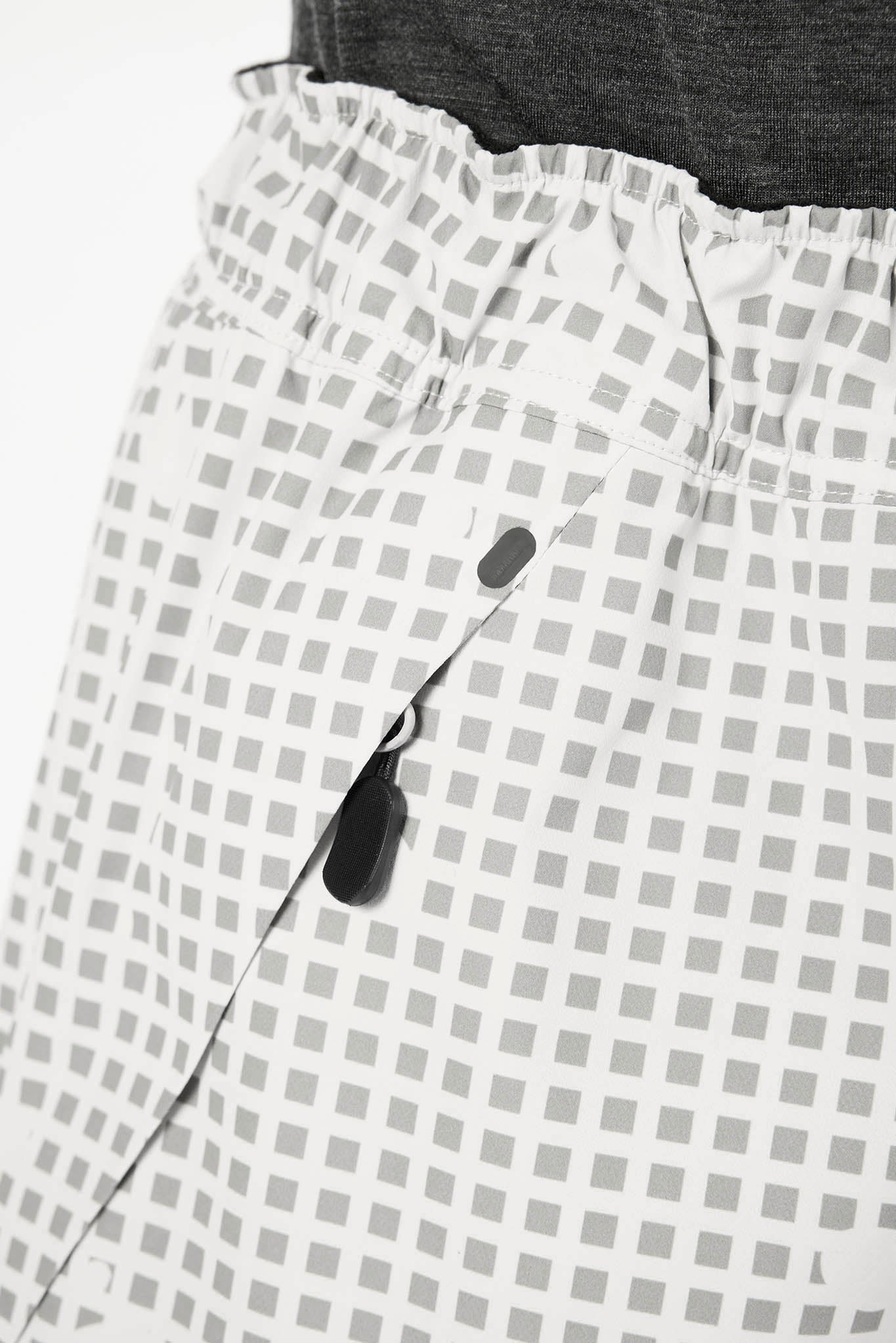Close-up of a white garment with a gray square grid pattern, featuring a black zipper pull and a dark gray waistband.