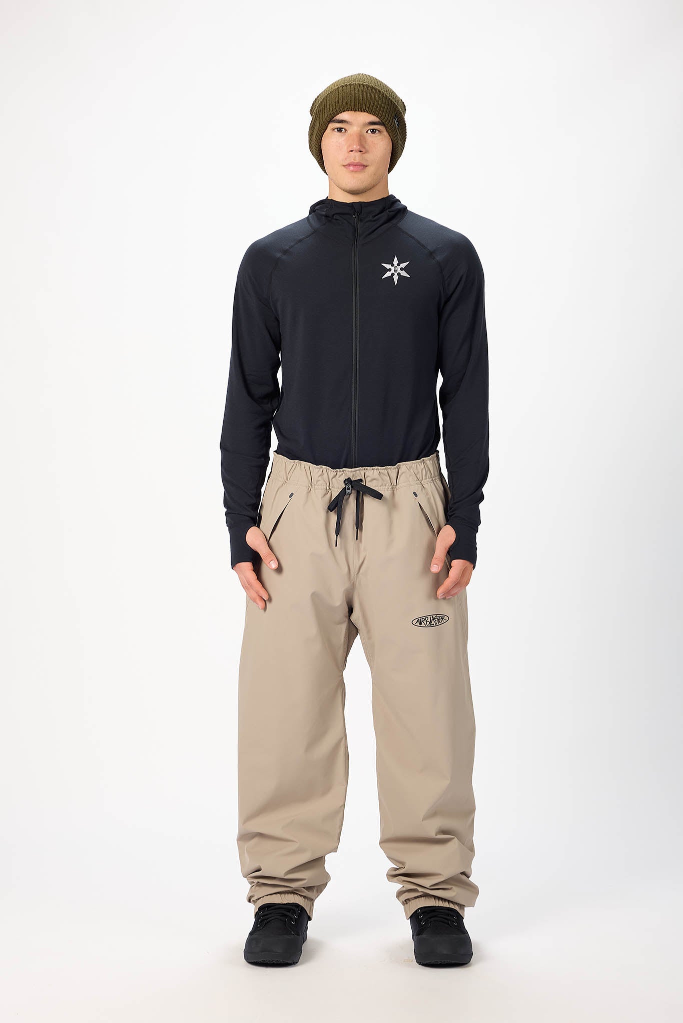 Person wearing a dark jacket with a logo, beige pants, and a green beanie on a white background