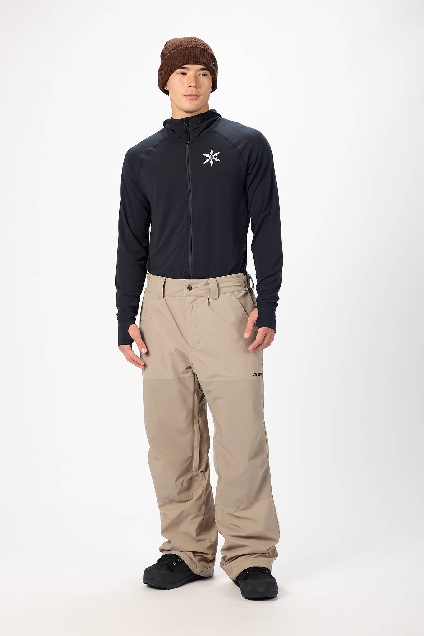 Person wearing a black jacket with a logo and beige pants on a white background