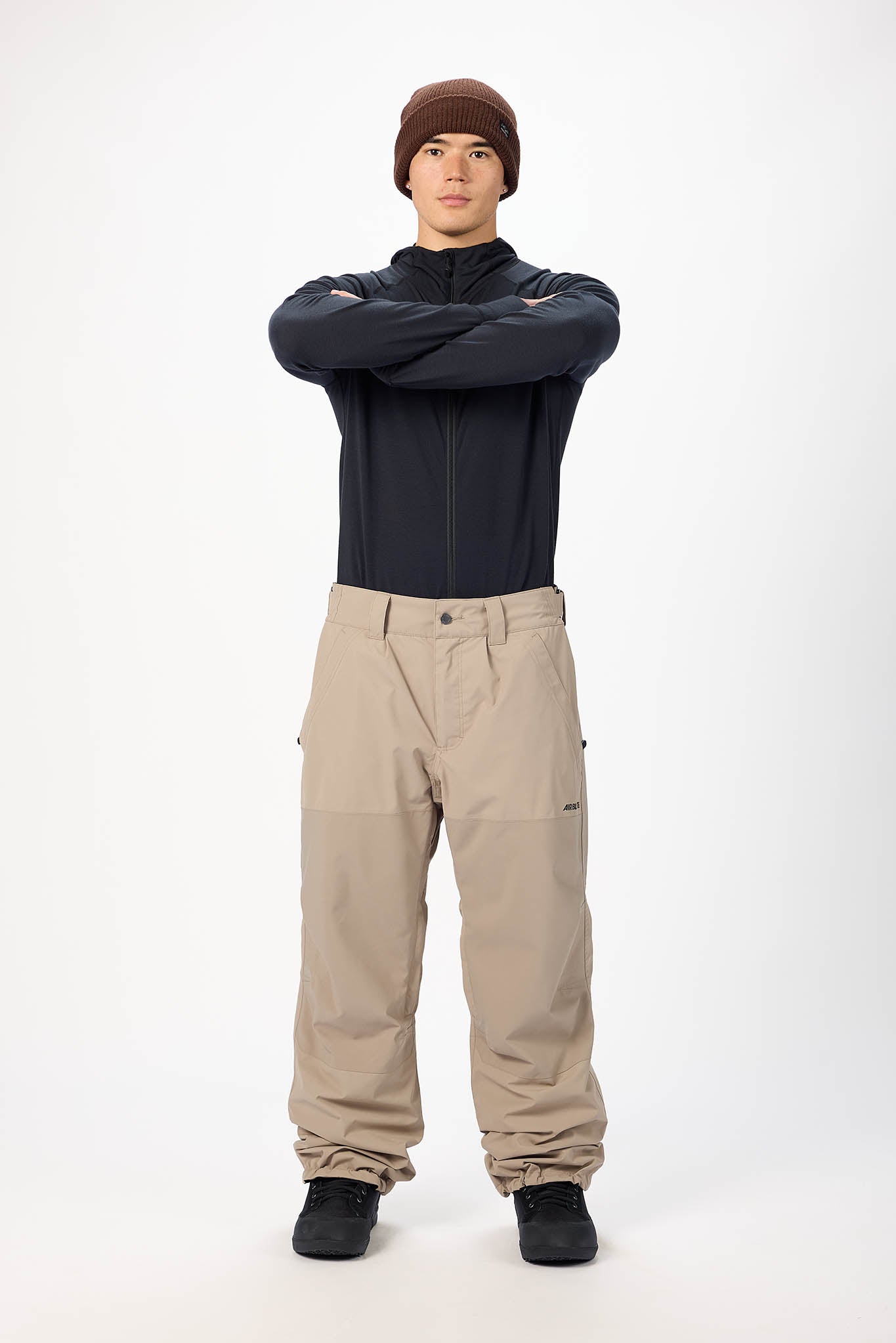 Person wearing a dark jacket, brown beanie, and beige pants on a white background