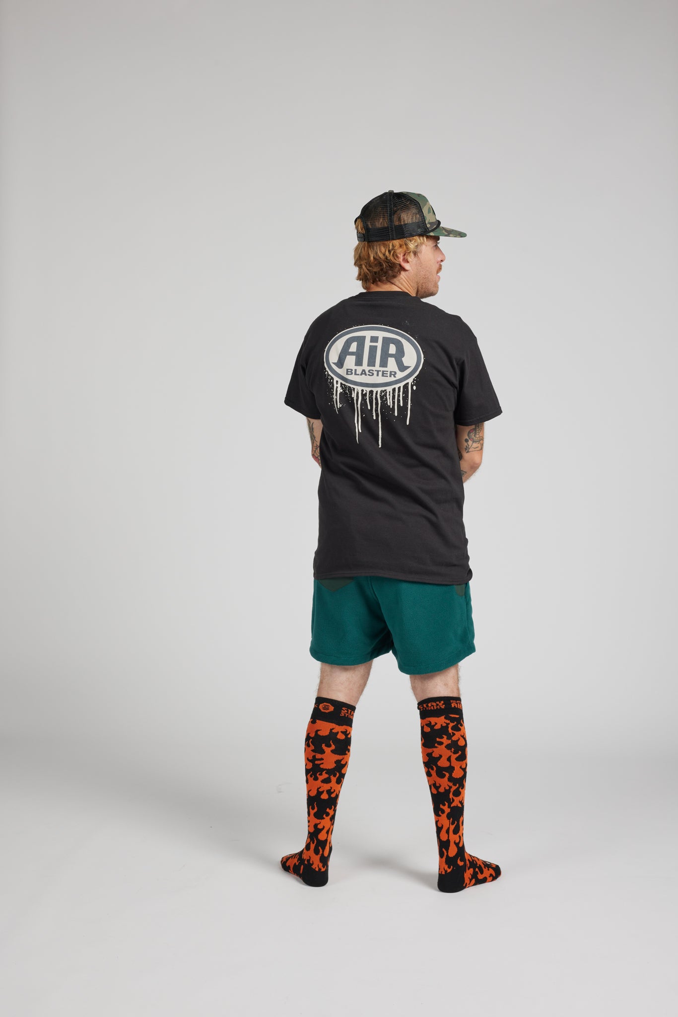 A model is shown from the back wearing a black t-shirt with a white 'AIR' logo, green shorts, and orange-and-black patterned socks, standing against a light gray studio background.