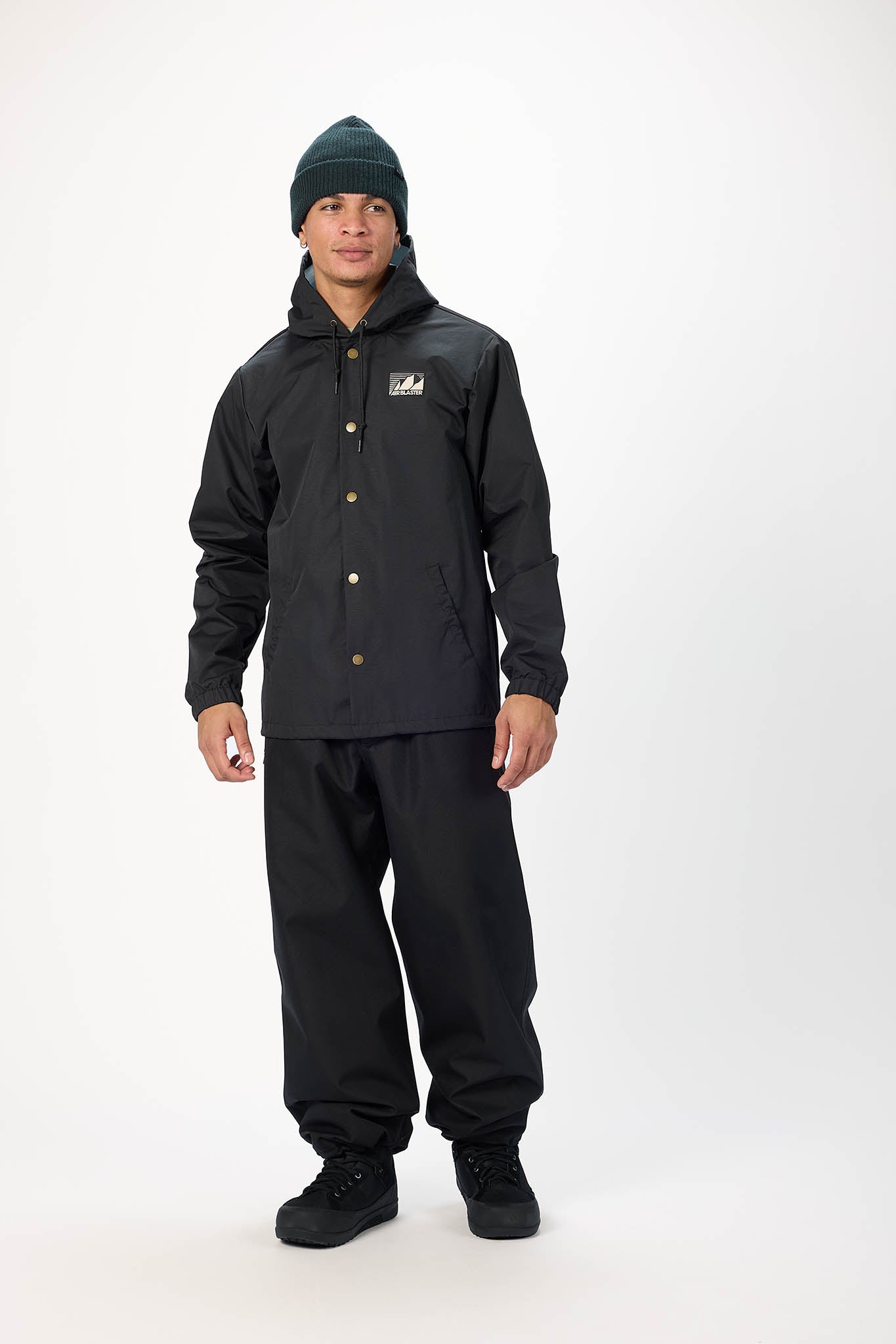 A man modeling a black hooded jacket and matching black pants, paired with a dark green beanie and black boots, against a plain white studio background.