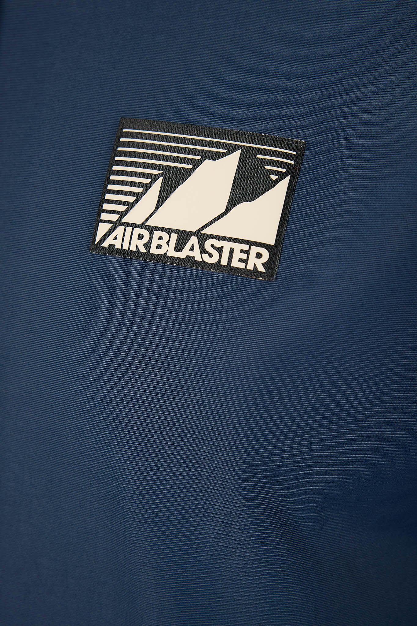 A close-up of a blue textured fabric featuring a rectangular black and white Air Blaster logo patch.