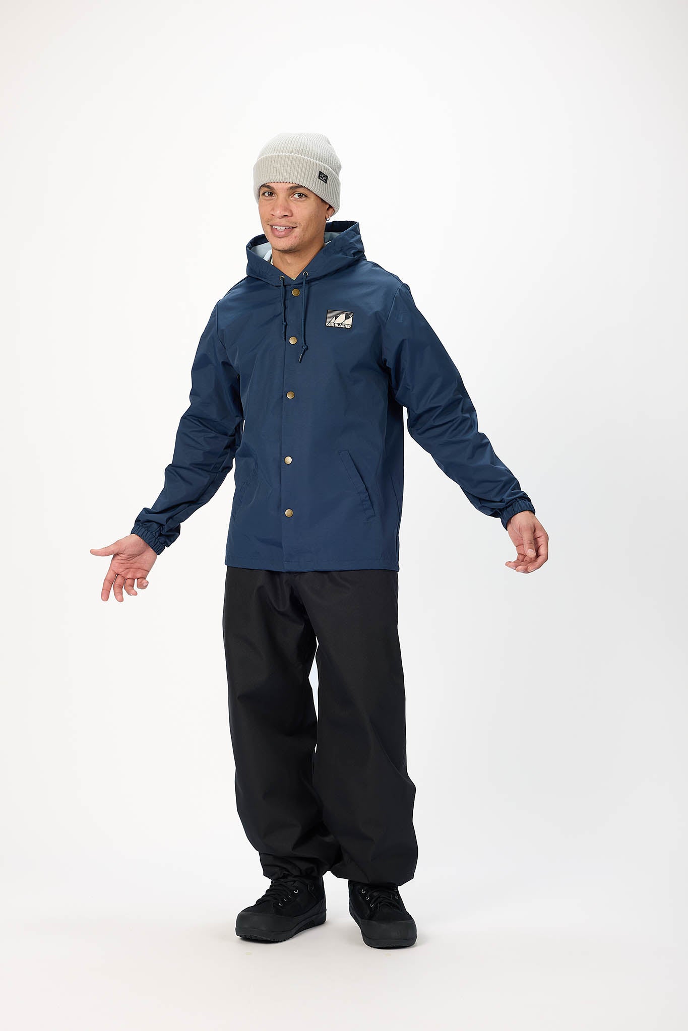 A man modeling a navy blue hooded jacket with snap buttons, black snow pants, and a light gray beanie against a plain white studio background.