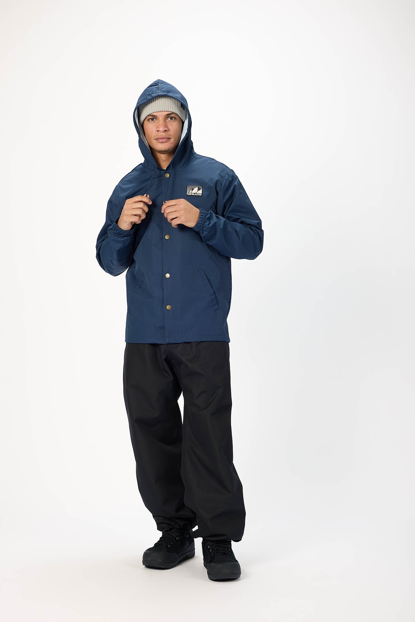 A man wearing a navy blue hooded jacket with gold buttons, black pants, and a gray beanie, standing against a plain white studio background.