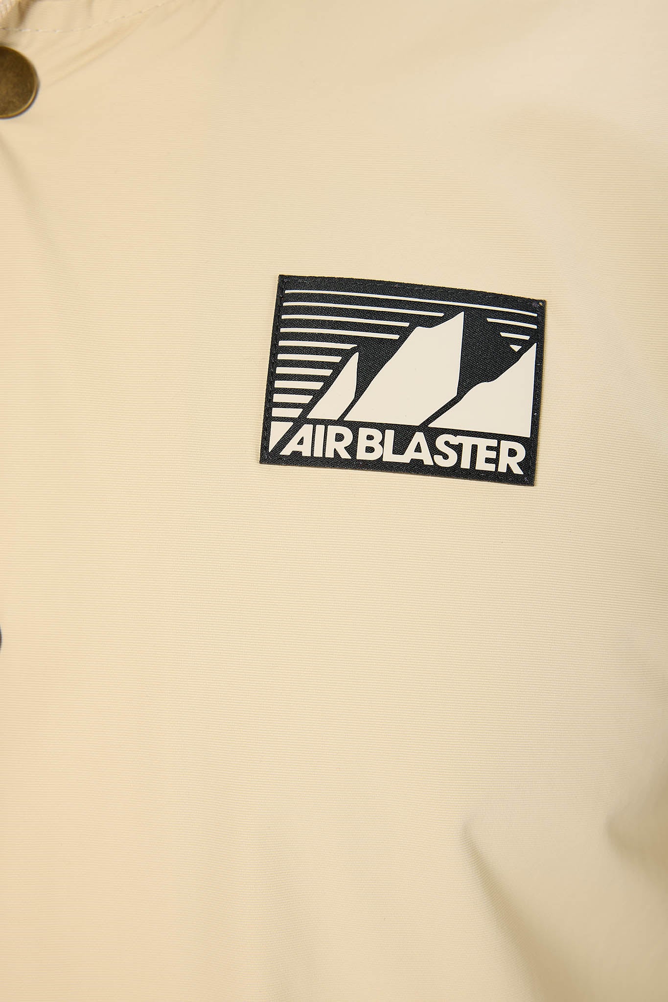 A close-up of a cream-colored garment featuring a black and white Airblaster logo patch on the chest.