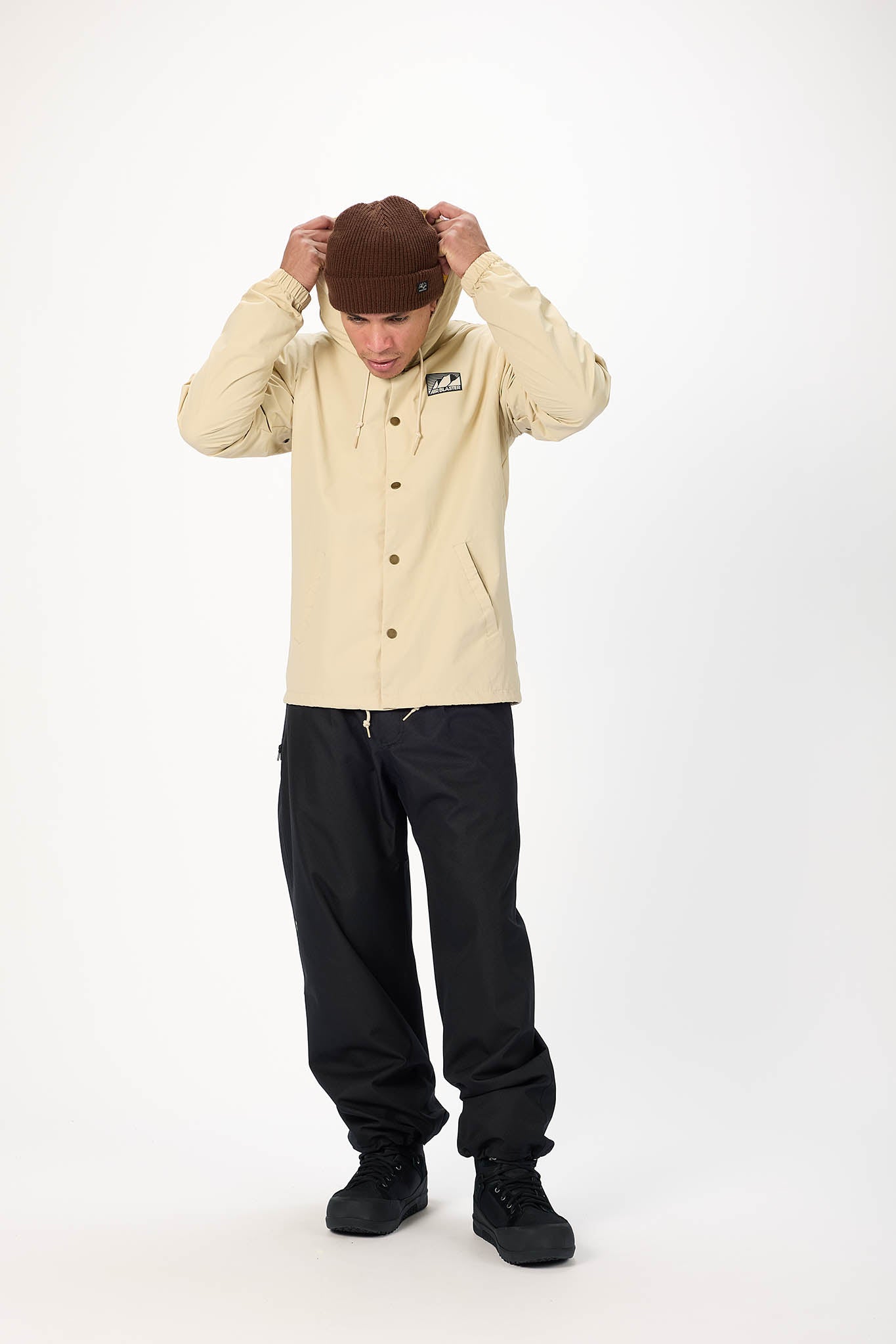 A model wearing a beige button-up jacket, black pants, and a brown beanie, standing against a plain white studio background.