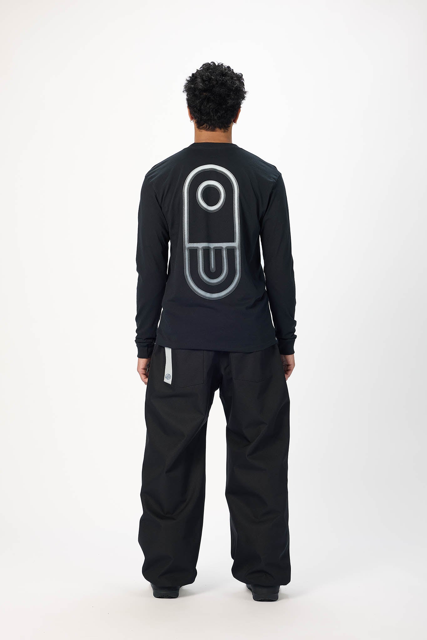 A model shown from the back wearing a black long-sleeve shirt with a large white outlined graphic on the back and black pants, photographed against a plain white studio background.