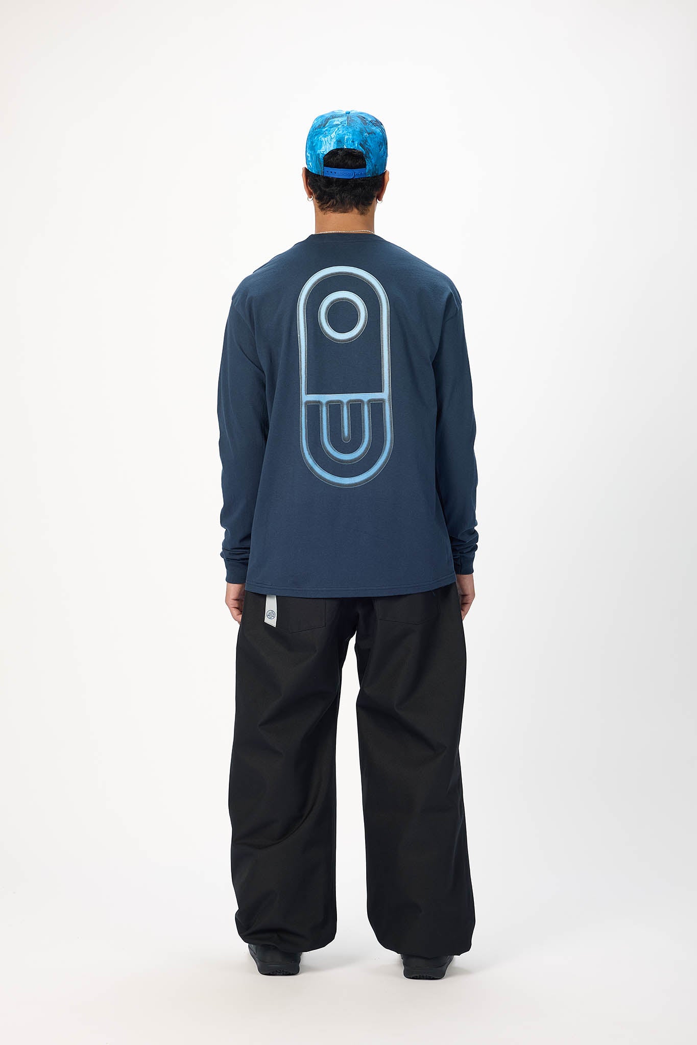 A model viewed from the back wearing a navy blue long-sleeve shirt with a large light blue graphic on the back, black pants, and a blue cap, against a plain white studio background.