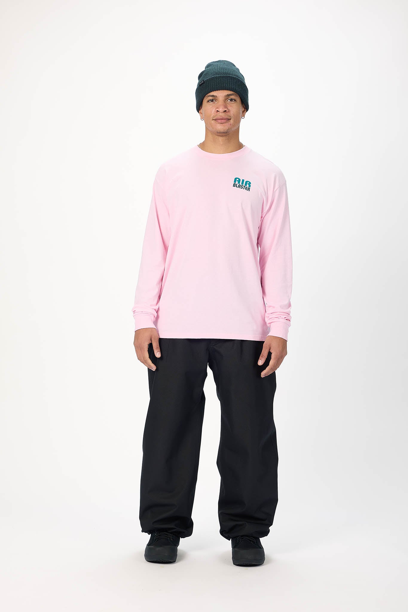 A model wearing a light pink long-sleeve t-shirt with a small teal graphic on the left chest, paired with black pants and a dark green beanie, standing against a plain white studio background.