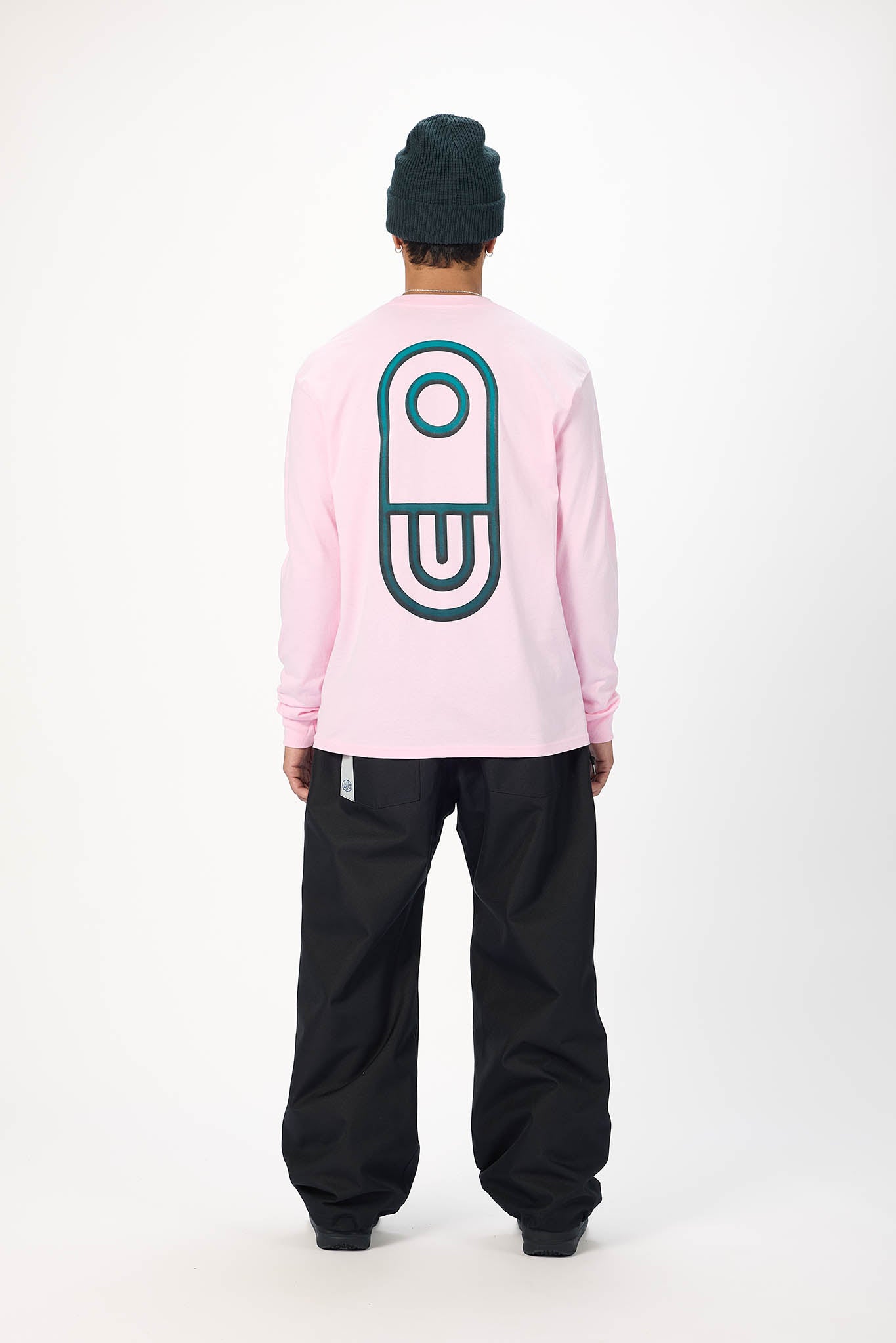 A person shown from the back wearing a light pink long-sleeve shirt with a large dark teal graphic on the back, black pants, and a black beanie, standing against a plain white studio background.