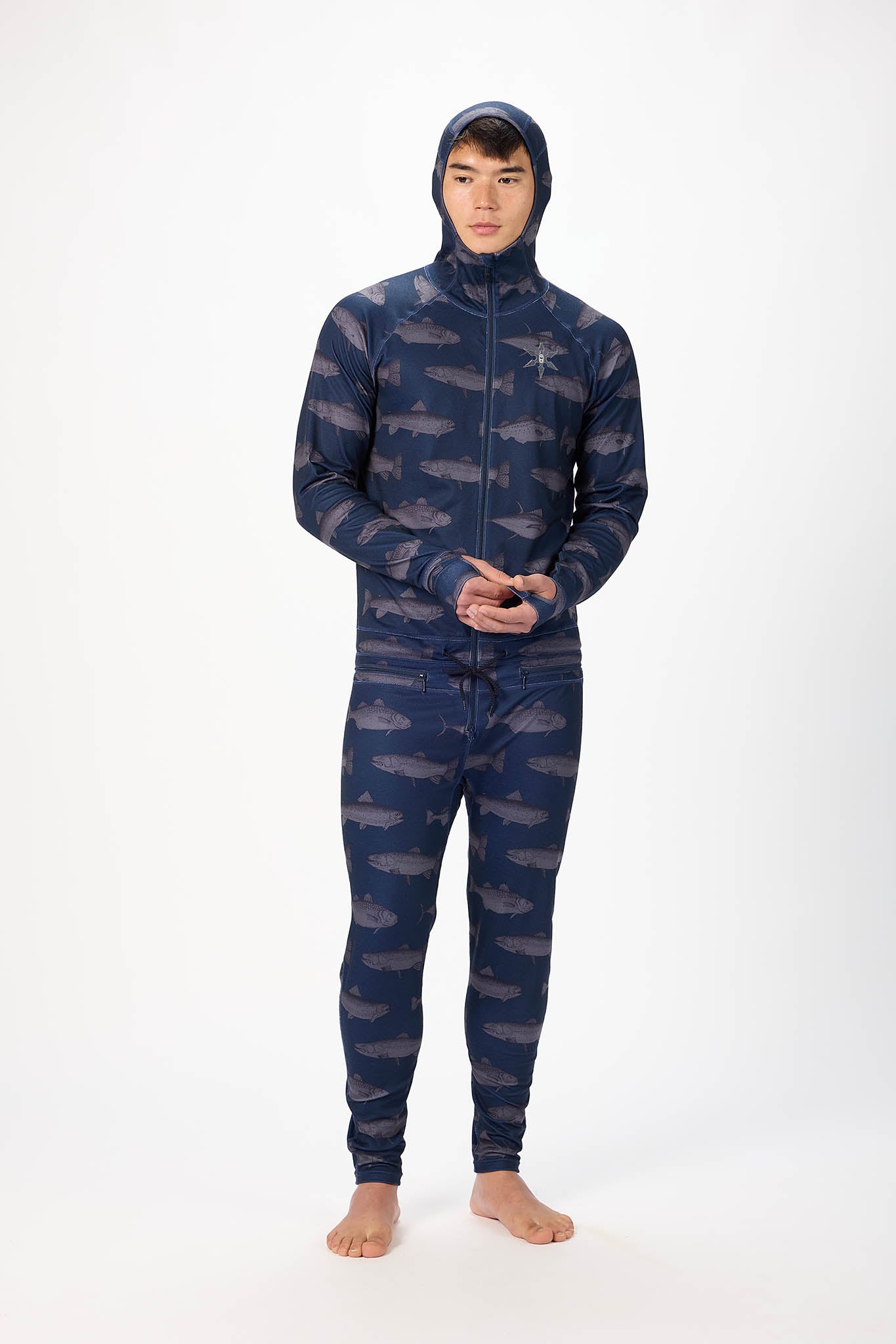 A man wearing a fish patterned onesie with a hooded with a zipper and jogger pants, standing against a plain white studio background.