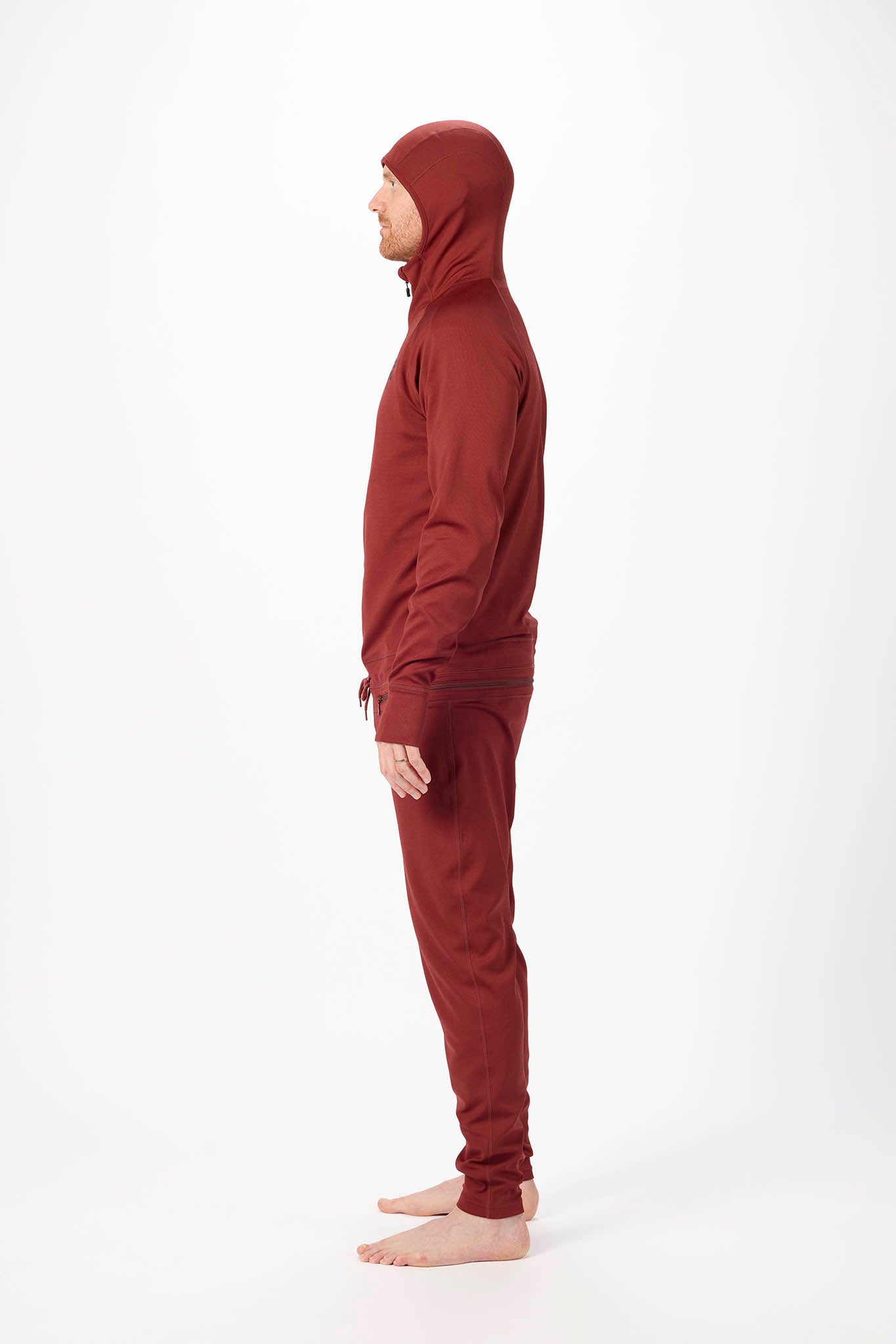 side profile A man wearing a matching burgundy onesie with a hooded zip-up jacket and jogger pants, standing against a plain white studio background.