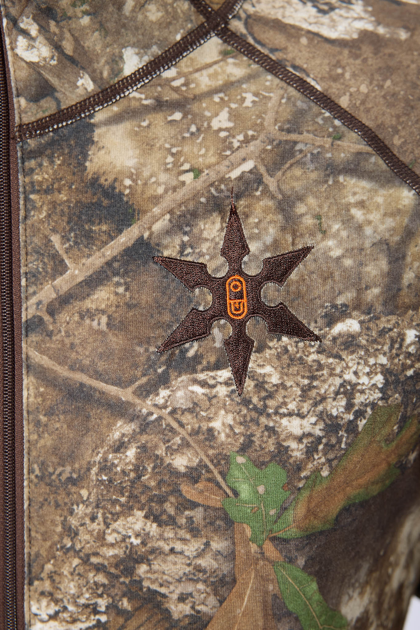 Close-up of a camouflage-patterned fabric with a dark brown six-pointed star patch featuring an orange 'GU' logo in the center.