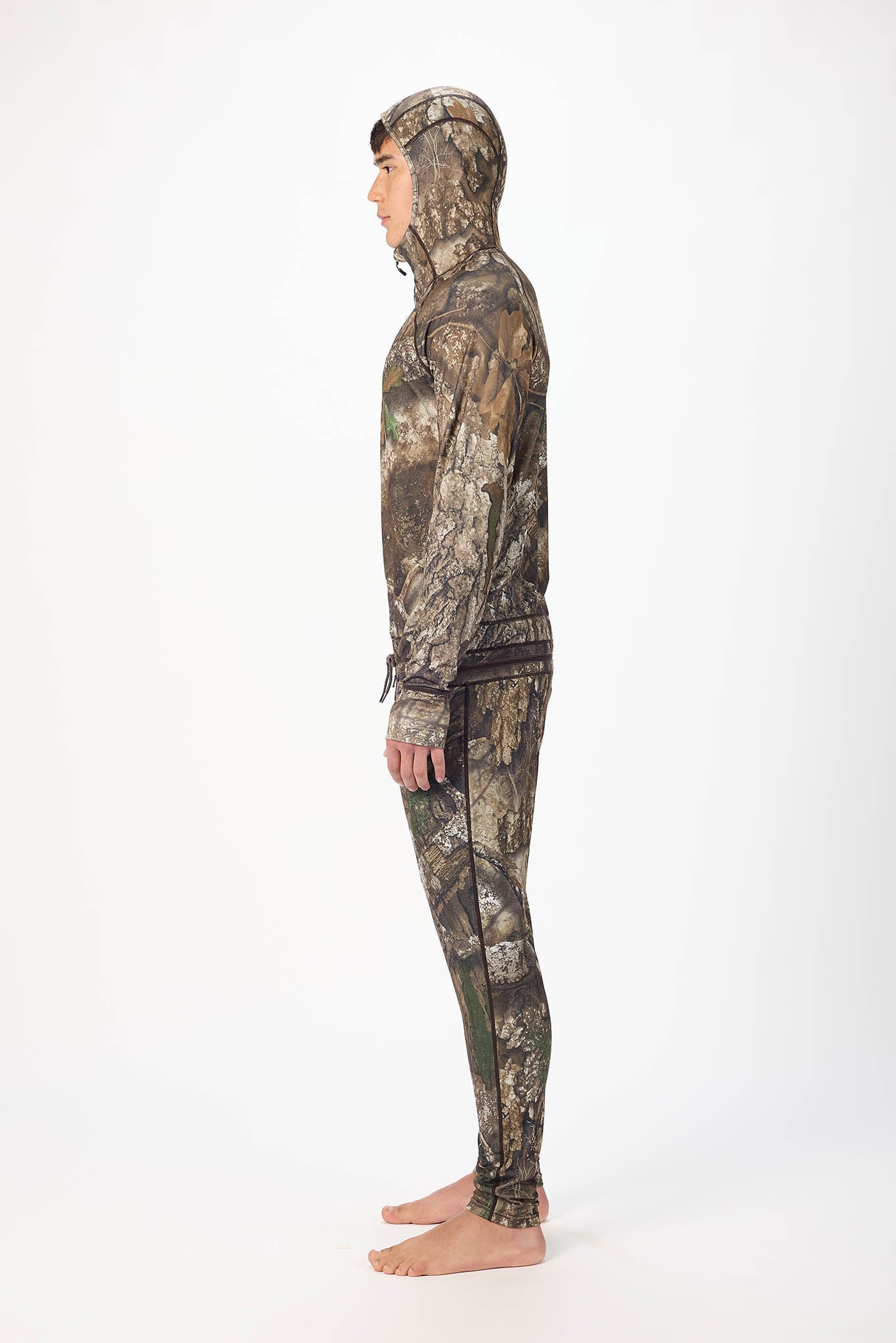 in side profile A man wearing a realtree camouflage  onesie with a hooded zip-up jacket and jogger pants, standing against a plain white studio background.