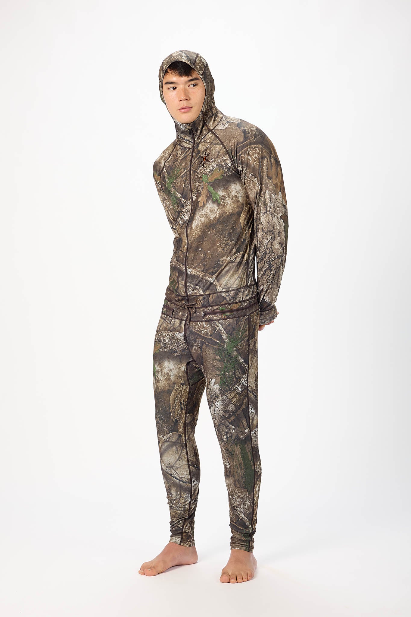 A man wearing a realtree camouflage  onesie with a hooded zip-up jacket and jogger pants, standing against a plain white studio background.