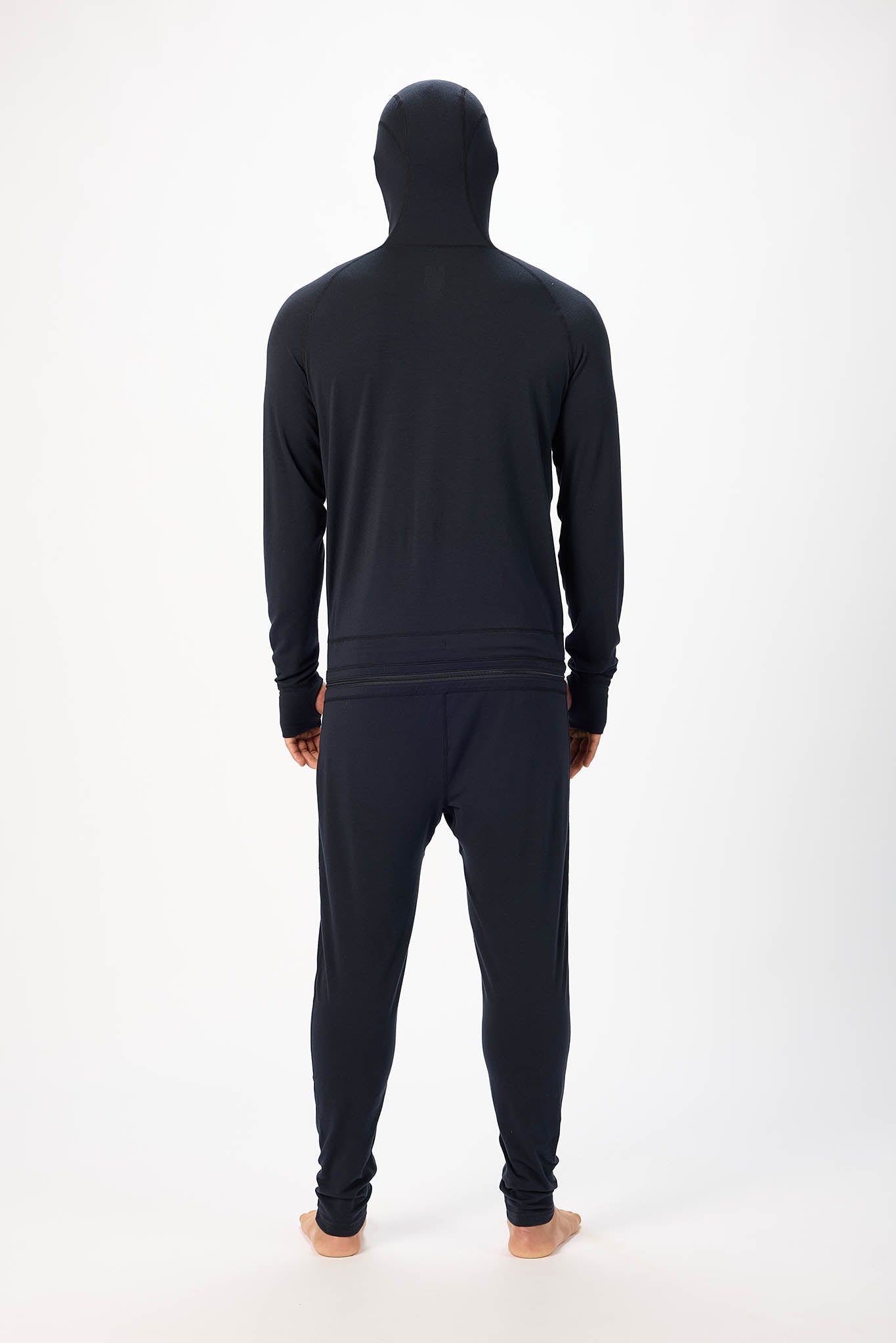 view from behind A man wearing a black onesie with a hooded zip-up and jogger pants, standing against a plain white studio background.
