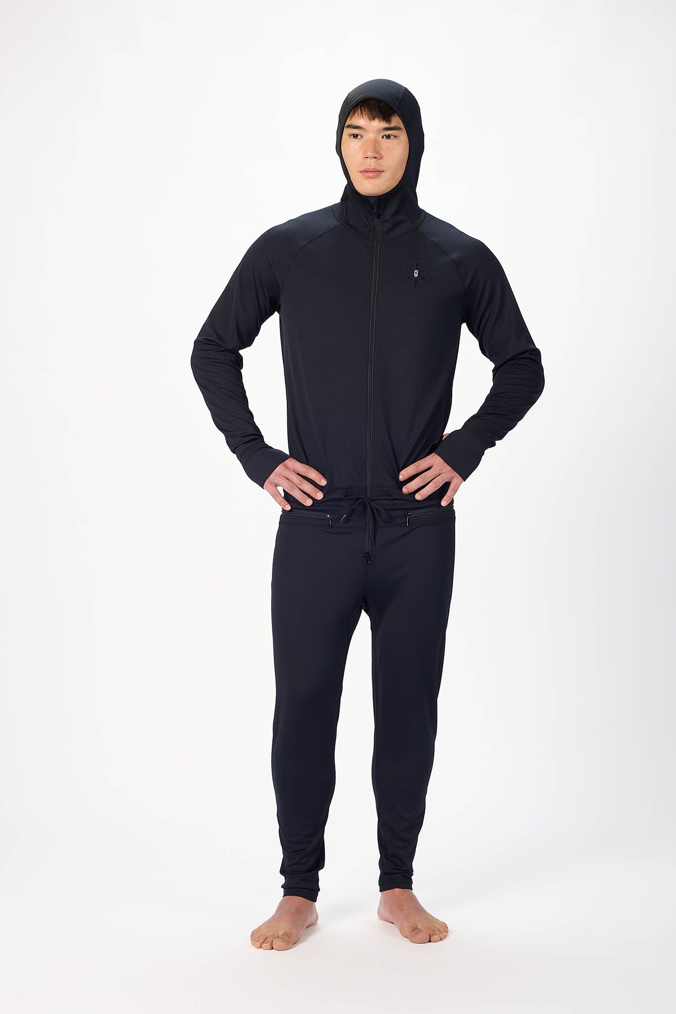 A man wearing a black onesie with a hooded with a zipper and jogger pants, standing against a plain white studio background.