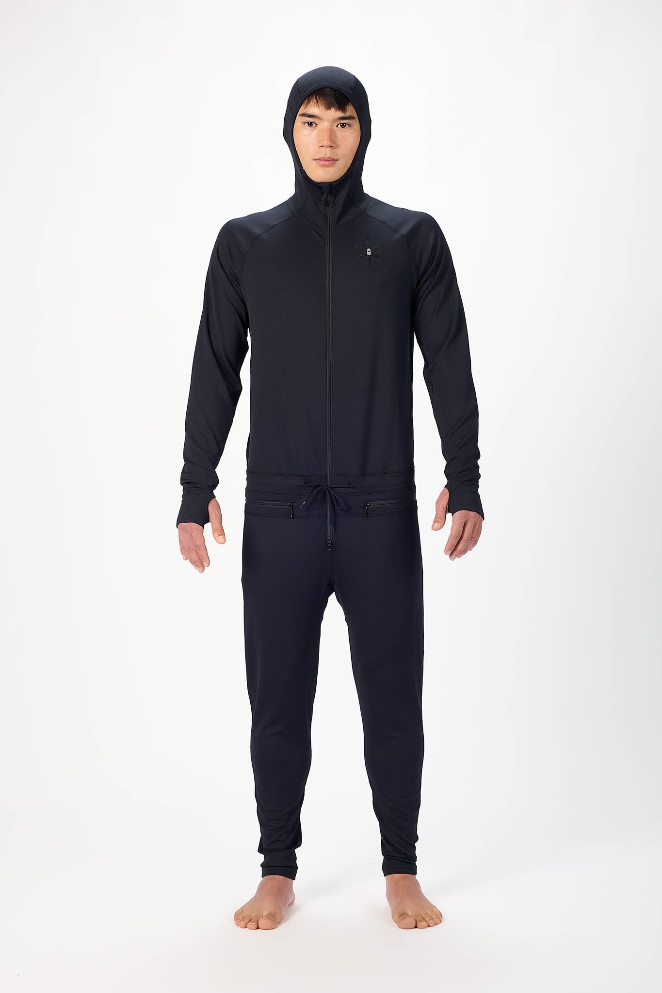 A man wearing a black  onesie with a hooded with a zipper and jogger pants, standing against a plain white studio background.