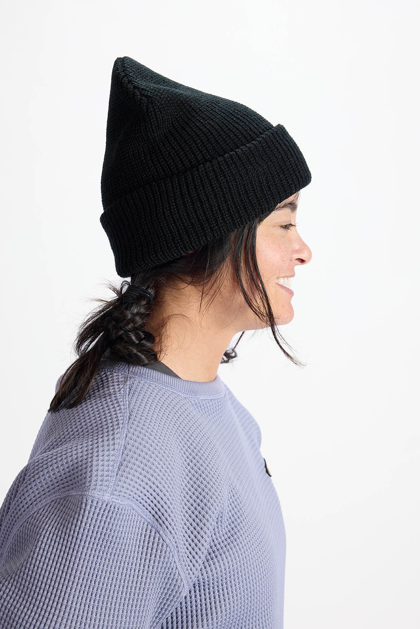 A woman in profile wearing a black ribbed knit beanie and a textured lavender-gray sweatshirt, photographed against a clean white background.