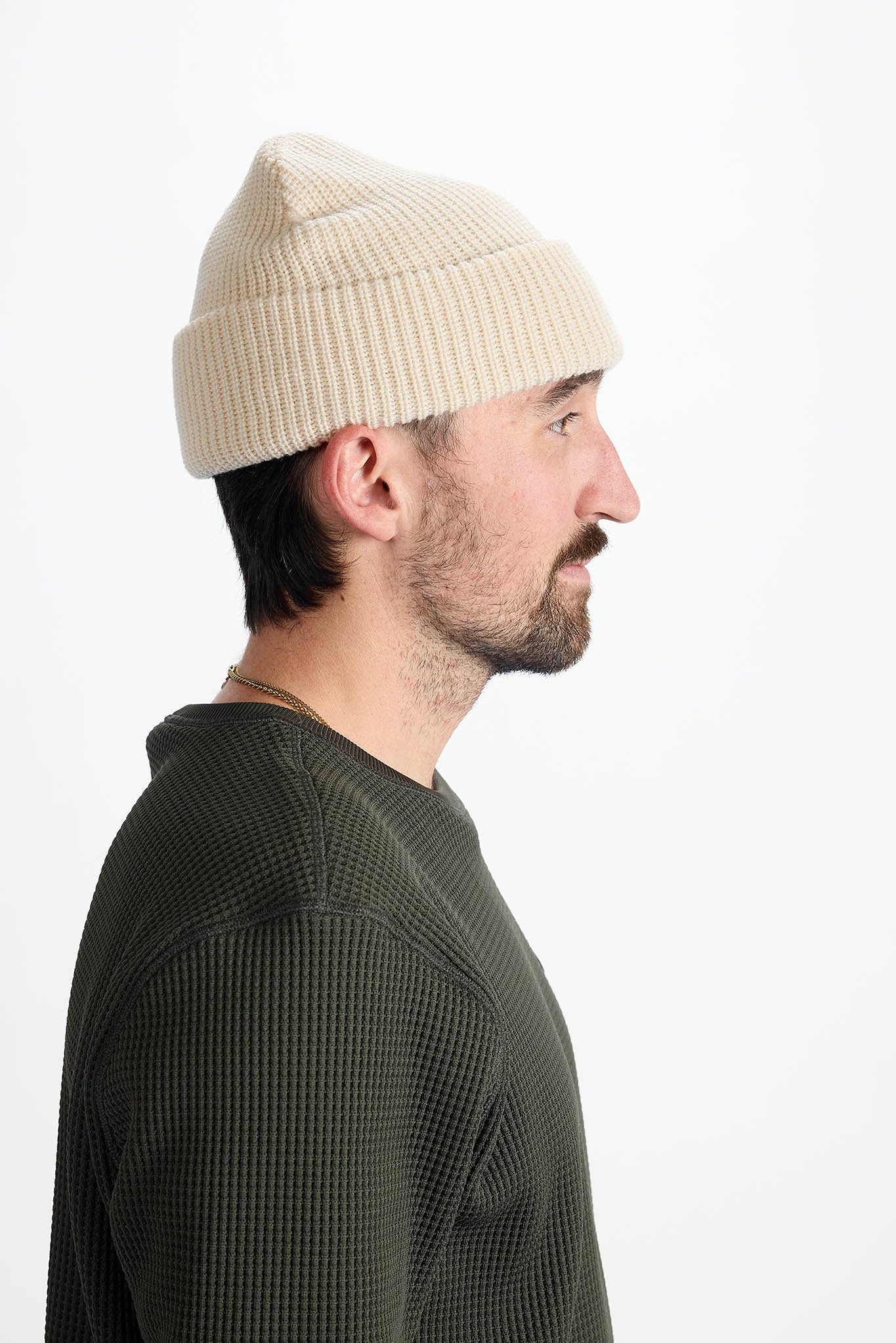 A man in profile wearing a cream-colored ribbed knit beanie and a dark olive green waffle-knit shirt, photographed against a clean white background.