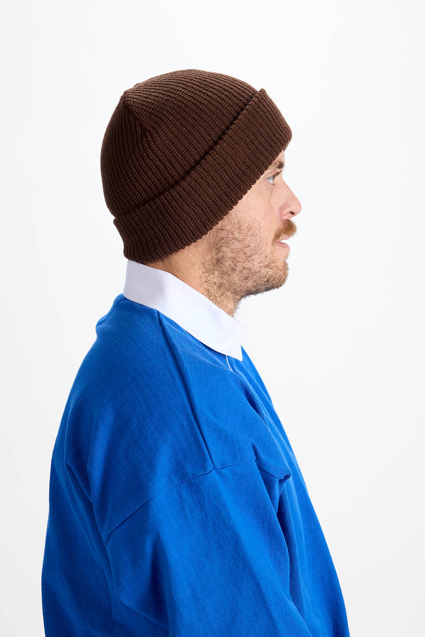 A man in profile wearing a dark brown ribbed knit beanie, a white collared shirt, and a blue sweater, photographed against a plain white studio background.