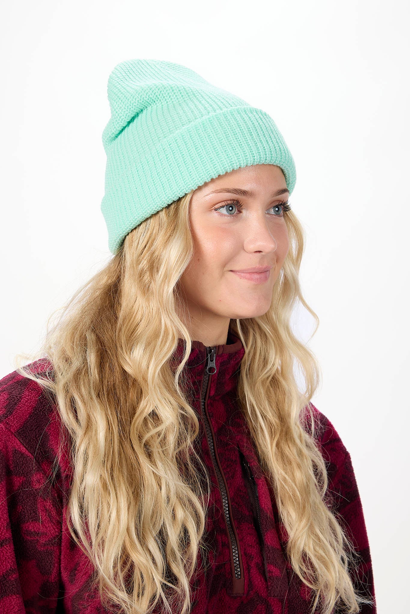 A woman with long blonde hair wearing a mint green ribbed knit beanie and a dark red patterned jacket, photographed against a plain white background.