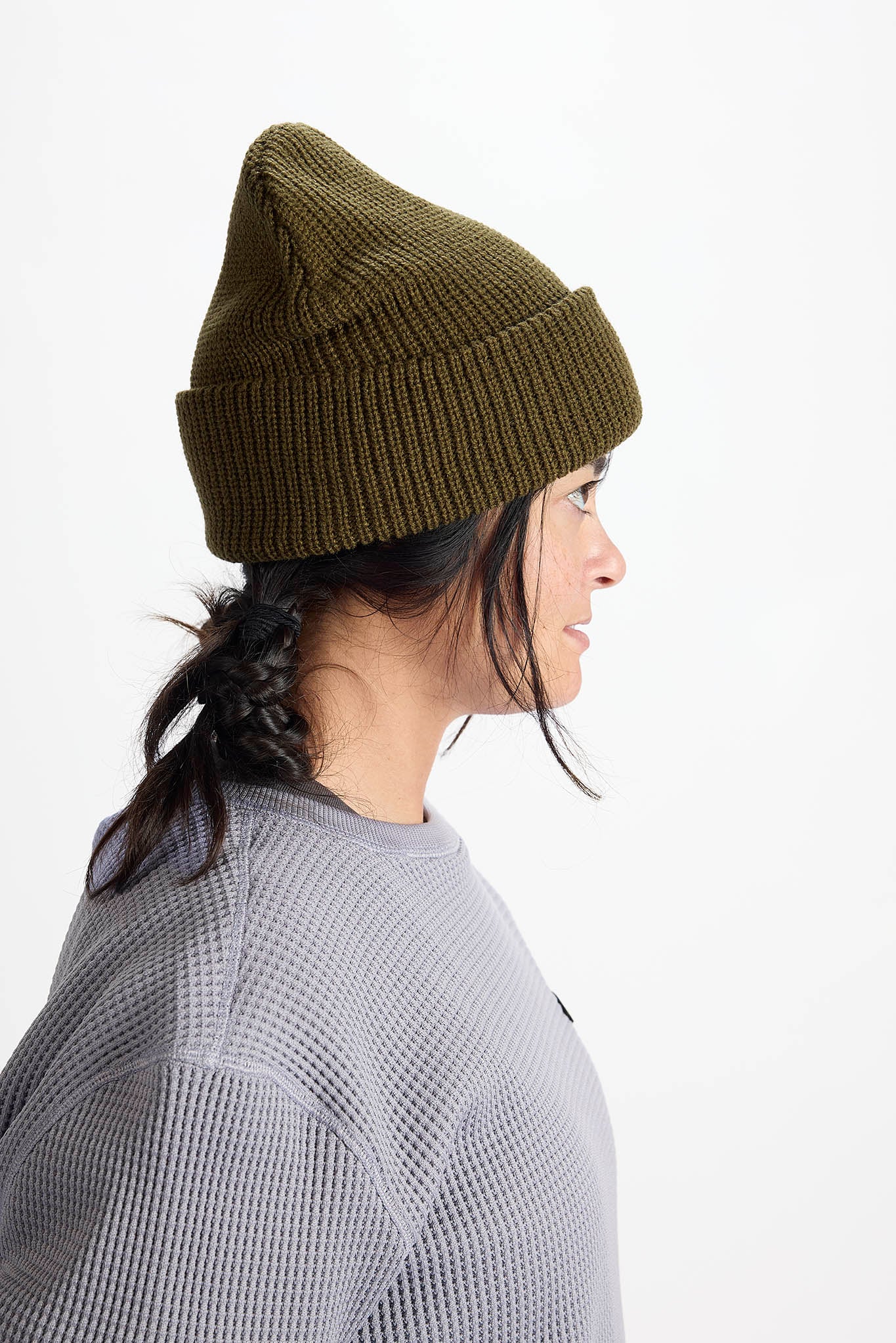 A woman shown in profile wearing an olive green ribbed knit beanie and a gray waffle-knit top against a plain white background.