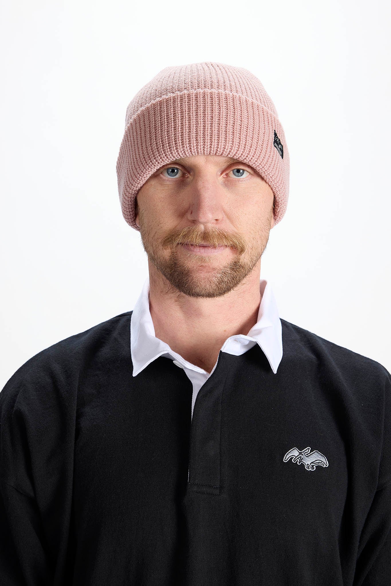 A man wearing a light pink ribbed knit beanie and a black rugby pullover over a white collared shirt, photographed against a plain white studio background.