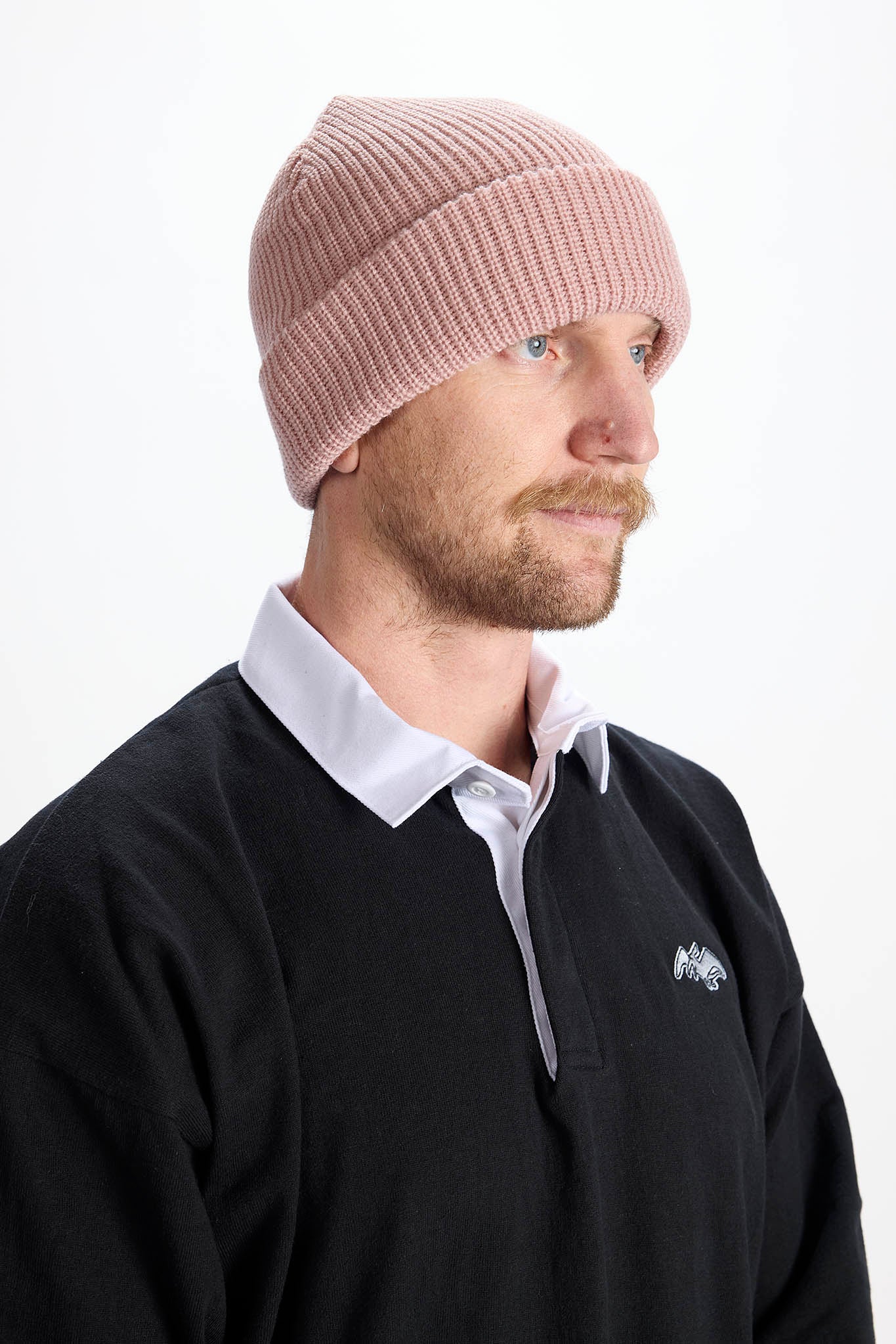 A man wearing a dusty pink ribbed knit beanie and a black collared pullover sweater, shown in profile against a plain white background.