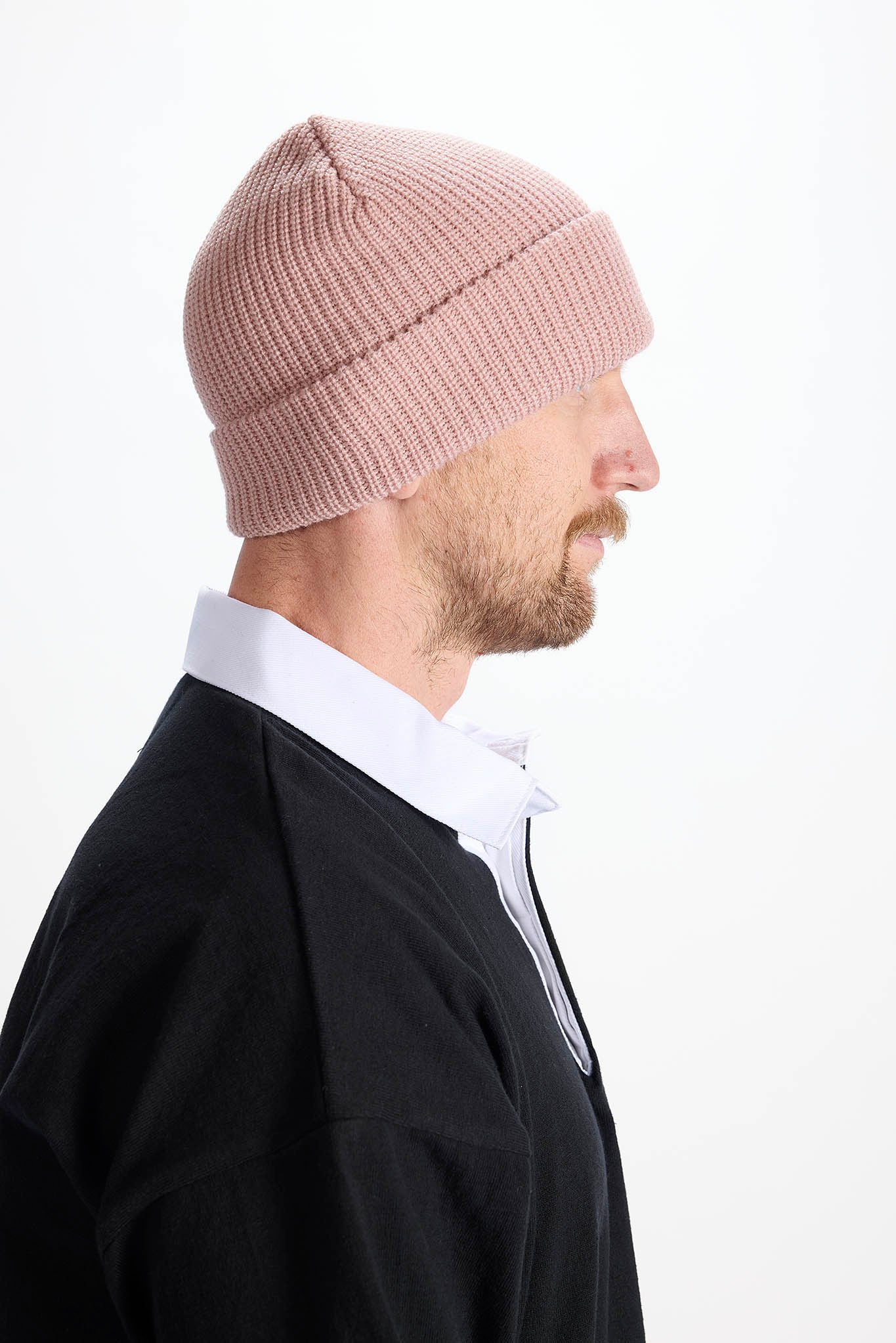 A man in profile wearing a light pink ribbed knit beanie, a white collared shirt, and a black sweater, photographed against a clean white background.